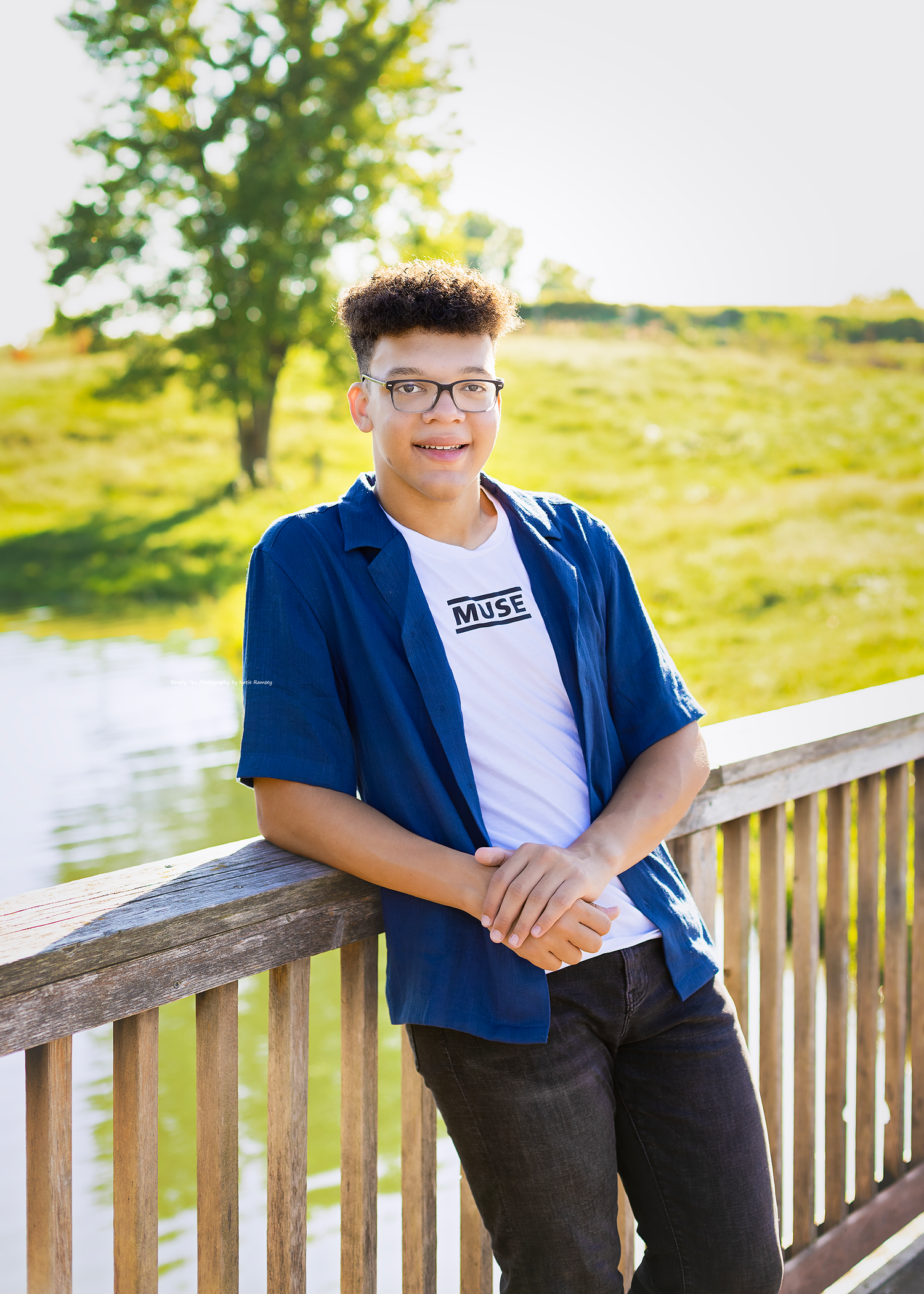 Teen boy in blue shirt and Muse tee leaning on wooden deck railing outdoors near pond.