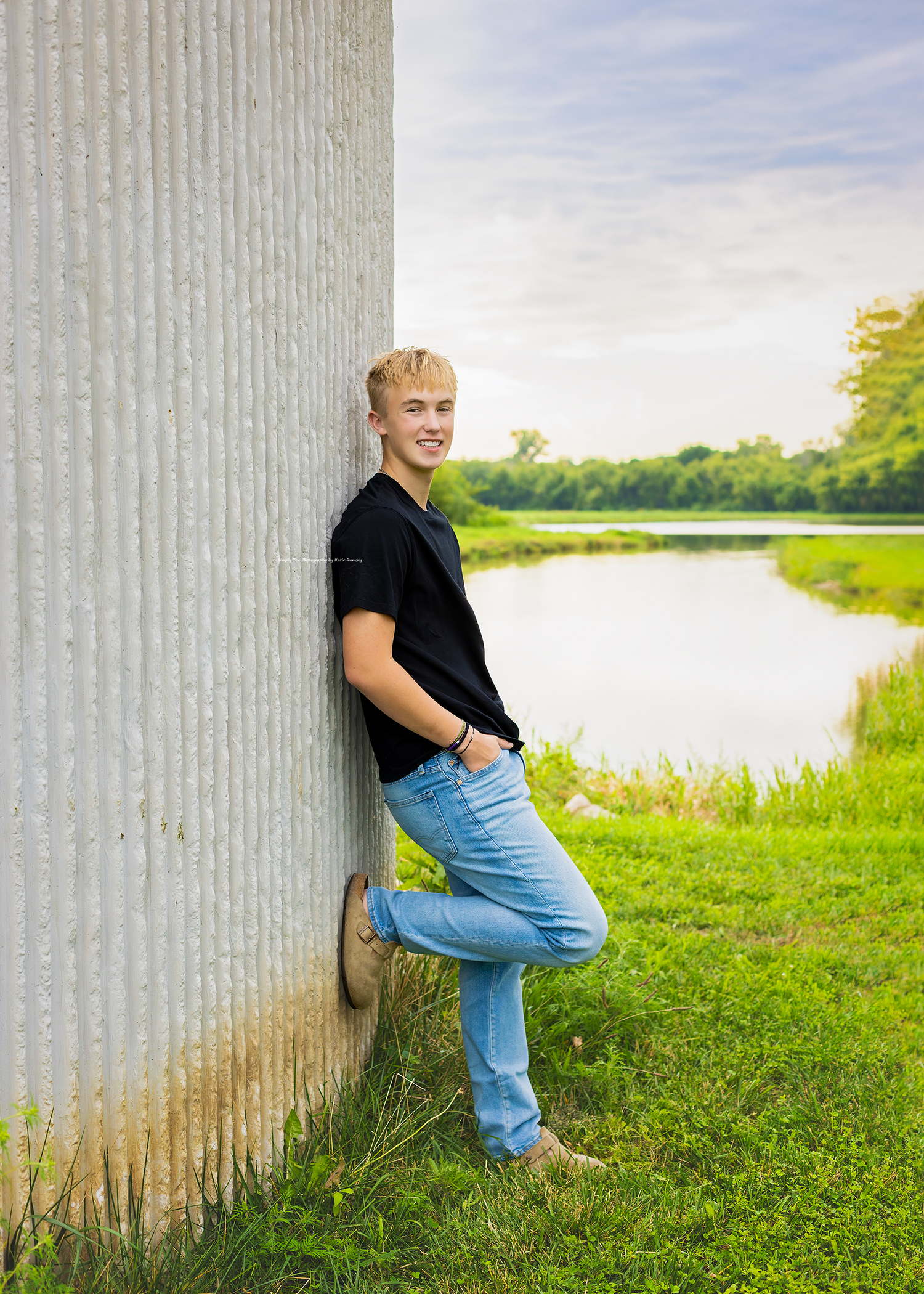 A smiling blonde teen boy leans against a metal grain bin wearing a black shirt and jeans near a pond.