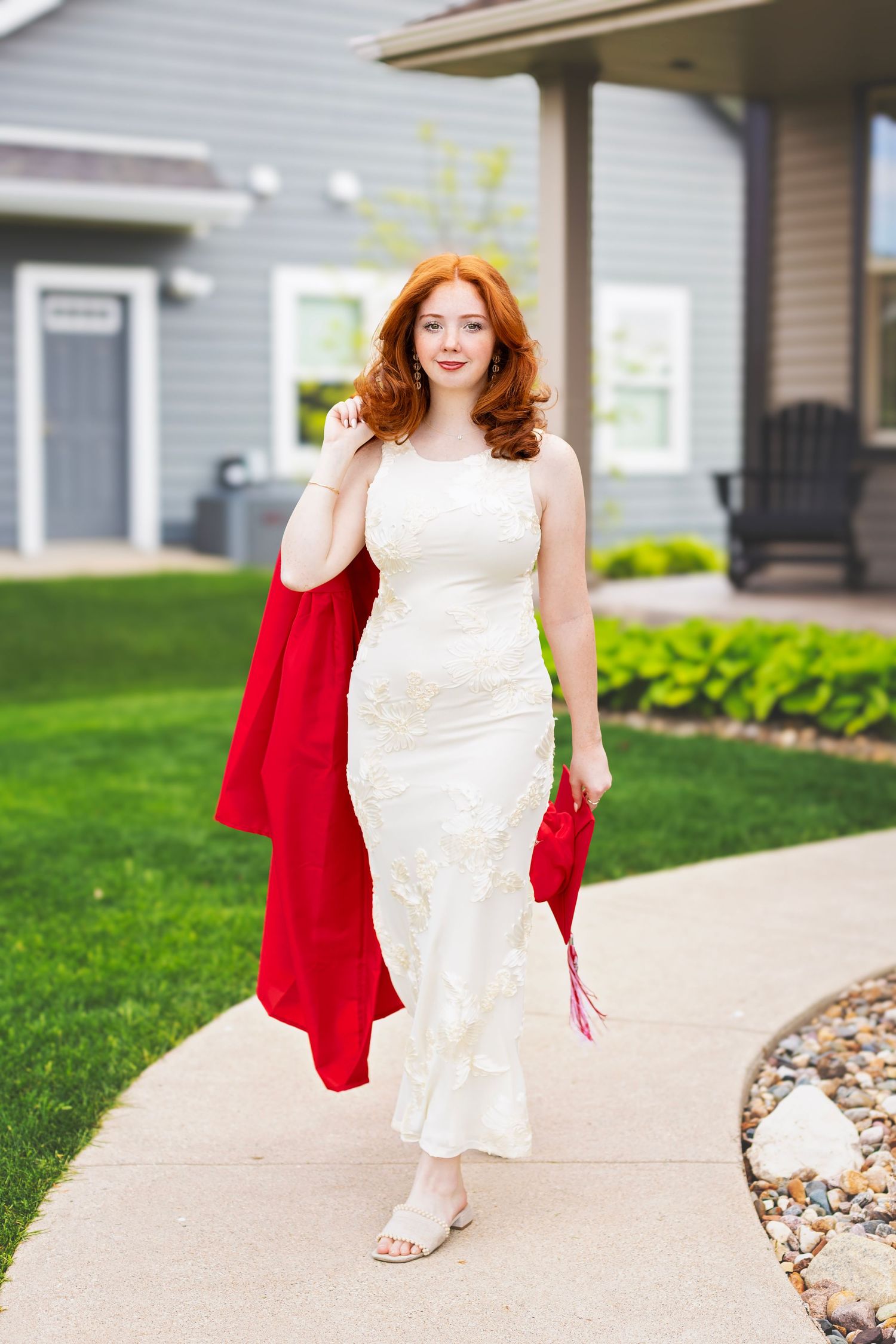 Red-haired young woman in white lace gown holding red graduation robe and cap on suburban sidewalk.