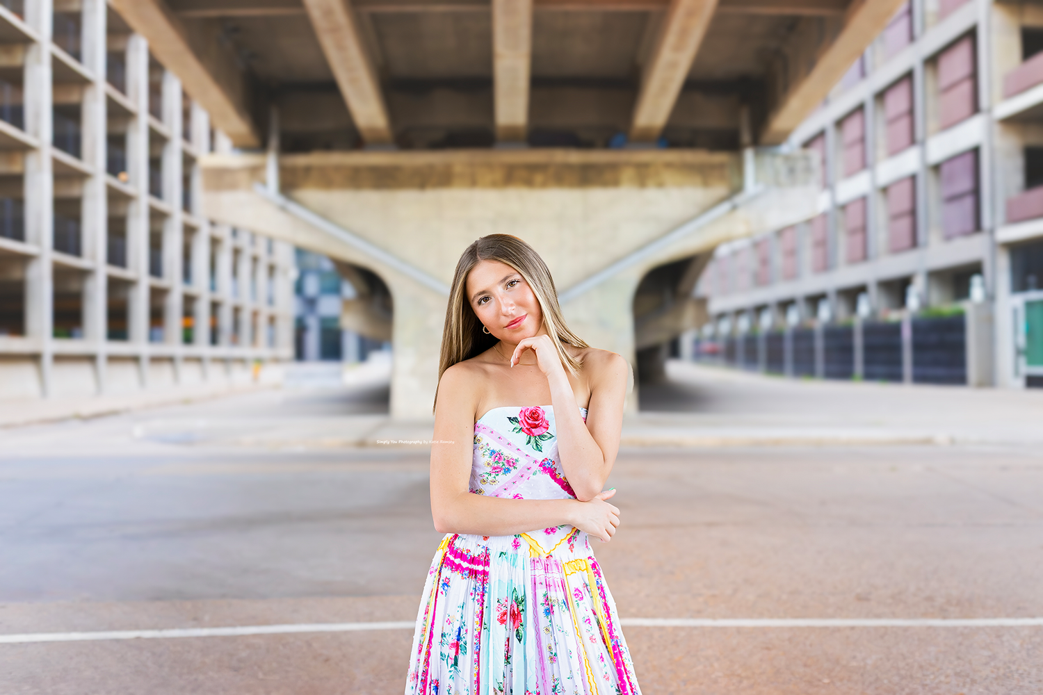 Young woman in colorful floral strapless dress posing thoughtfully under urban bridge architecture.