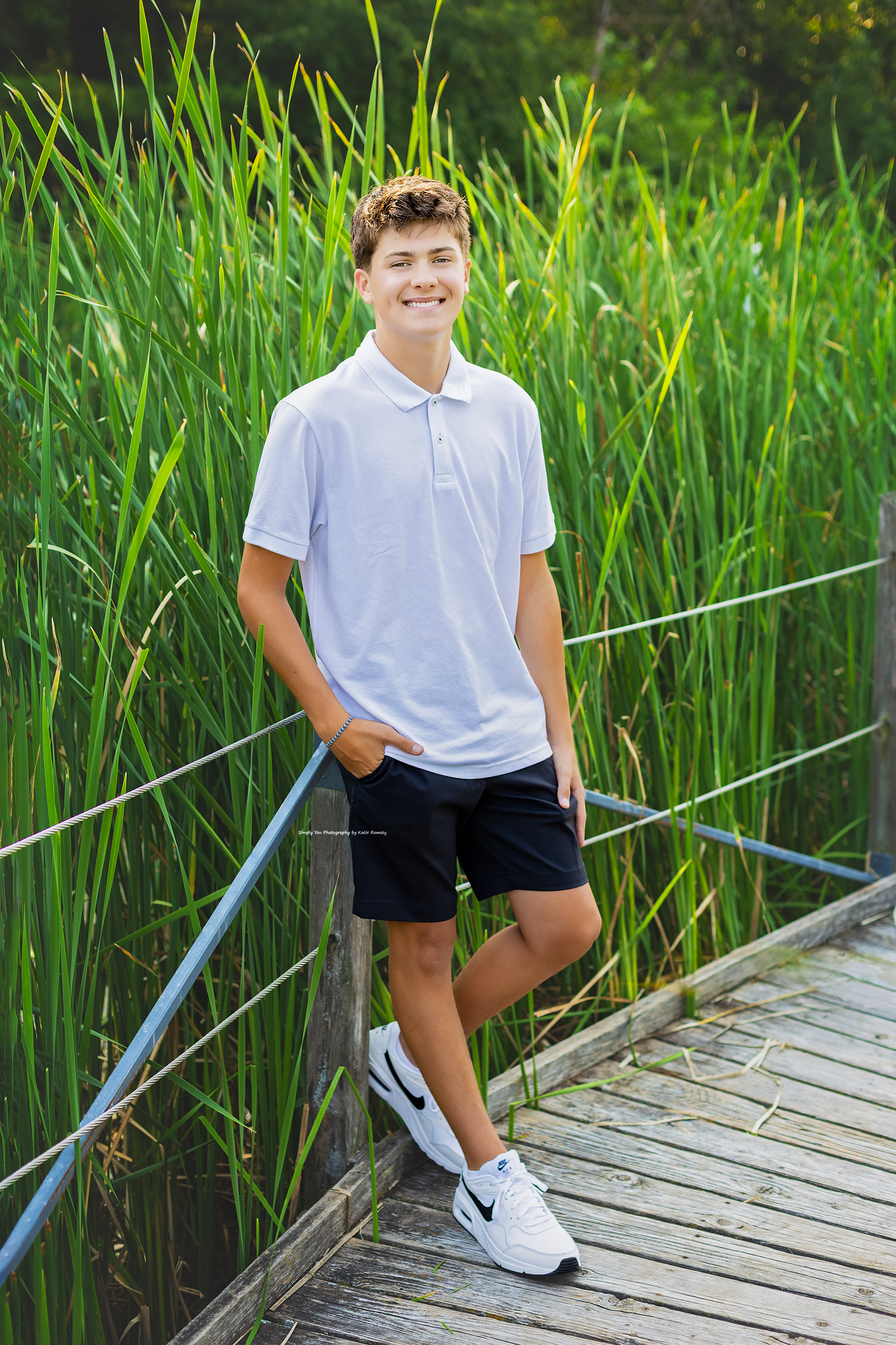 Smiling teenage boy in white polo shirt and black shorts standing on wooden dock surrounded by tall green reeds.