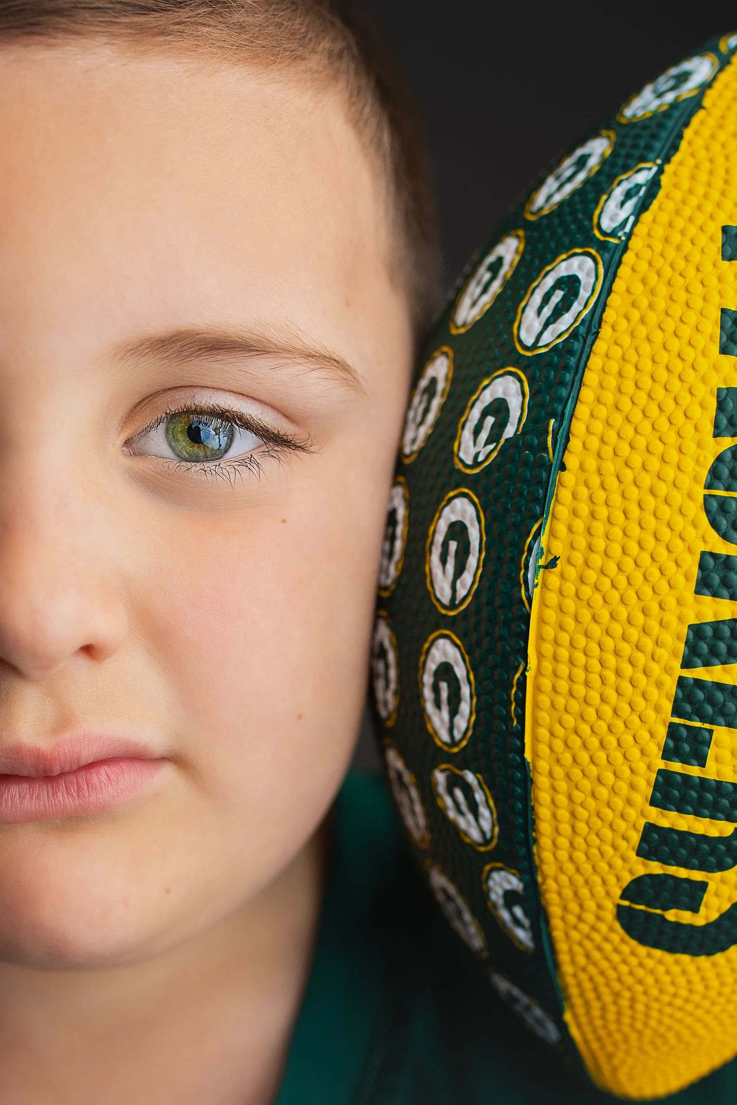 Close-up of child with blue eyes holding a Green Bay Packers football against dark background.