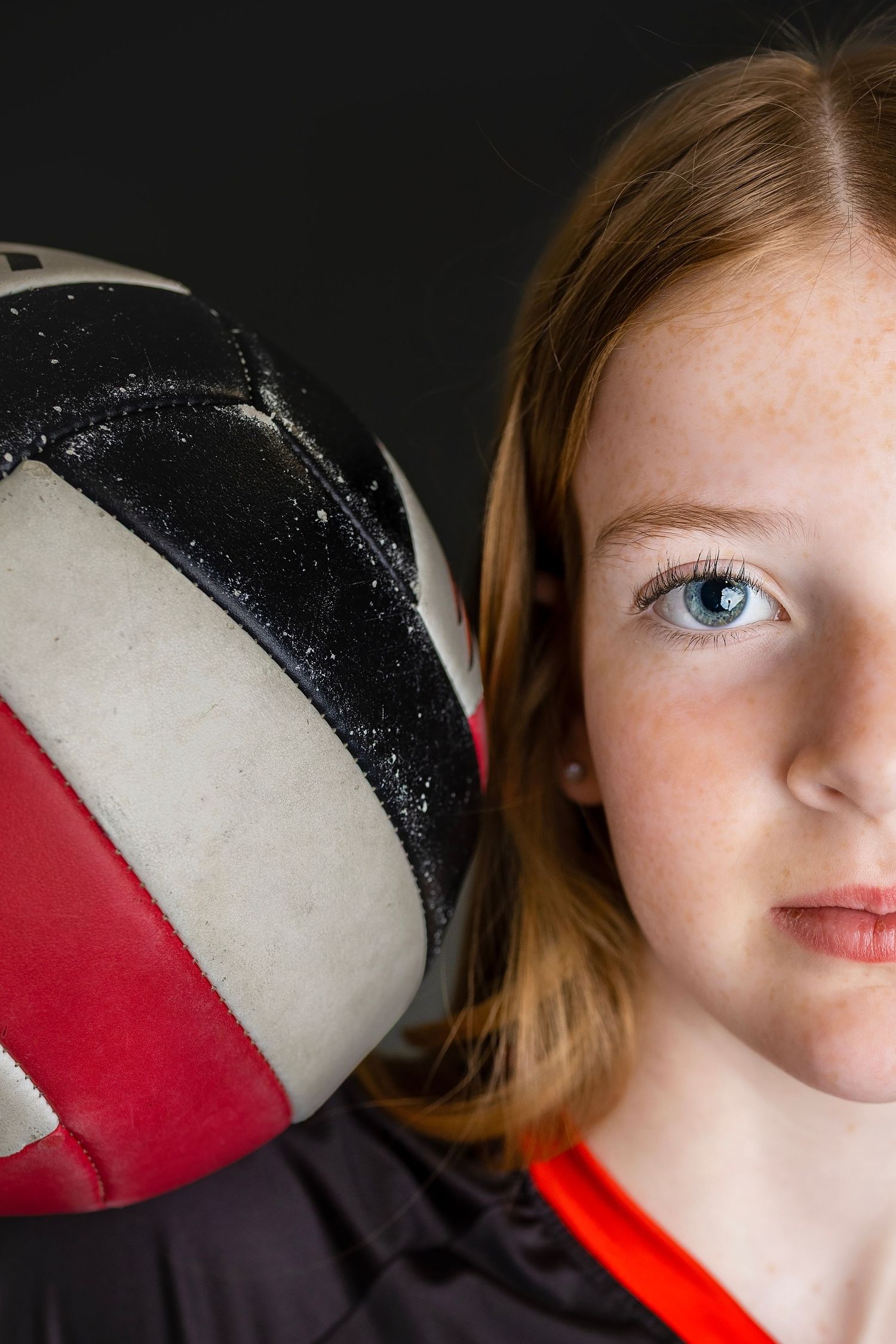 Close-up portrait of a young redheaded girl with blue eyes holding a black and red volleyball against her face.