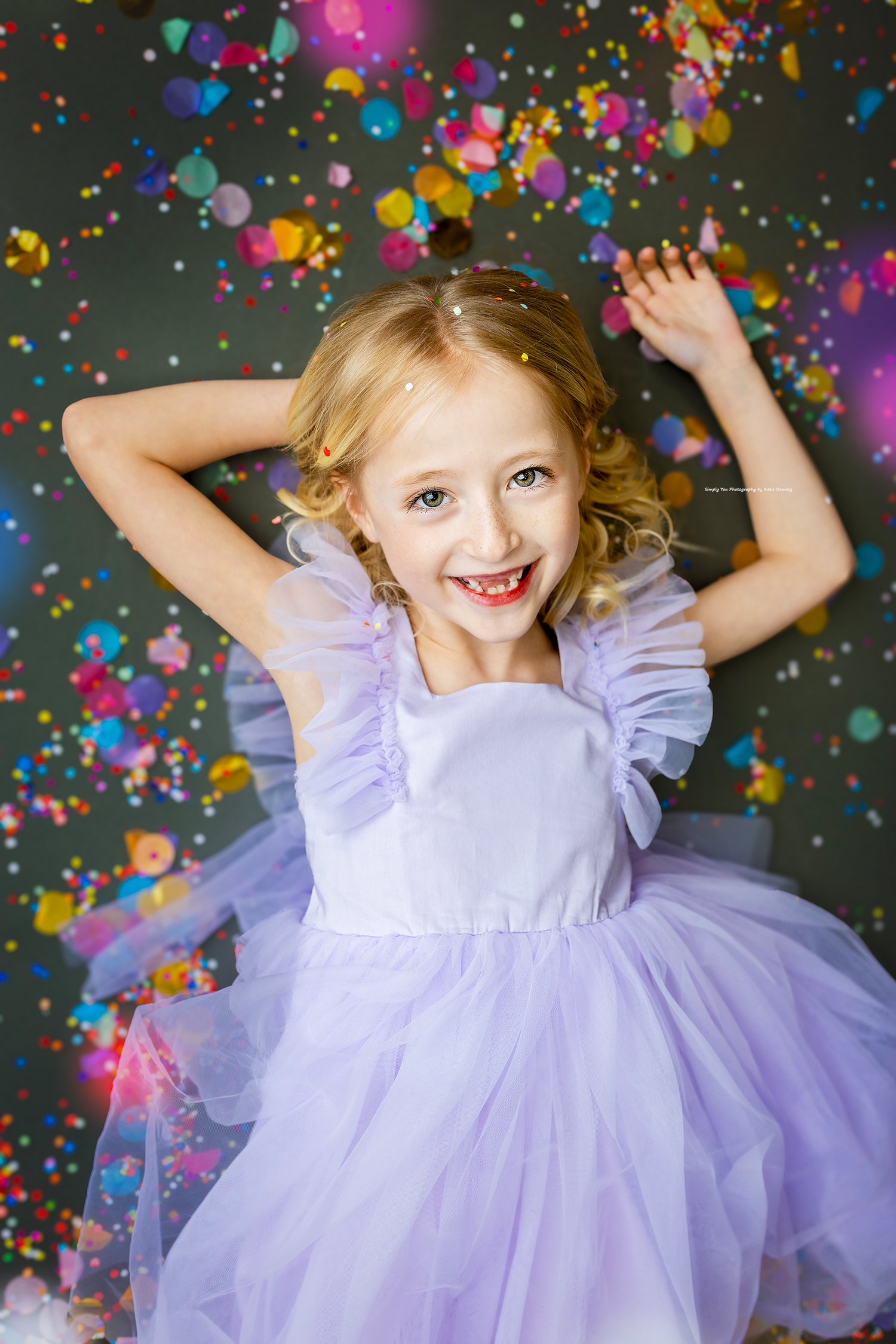 Joyful blonde girl in purple tulle dress dancing with colorful confetti falling around her.