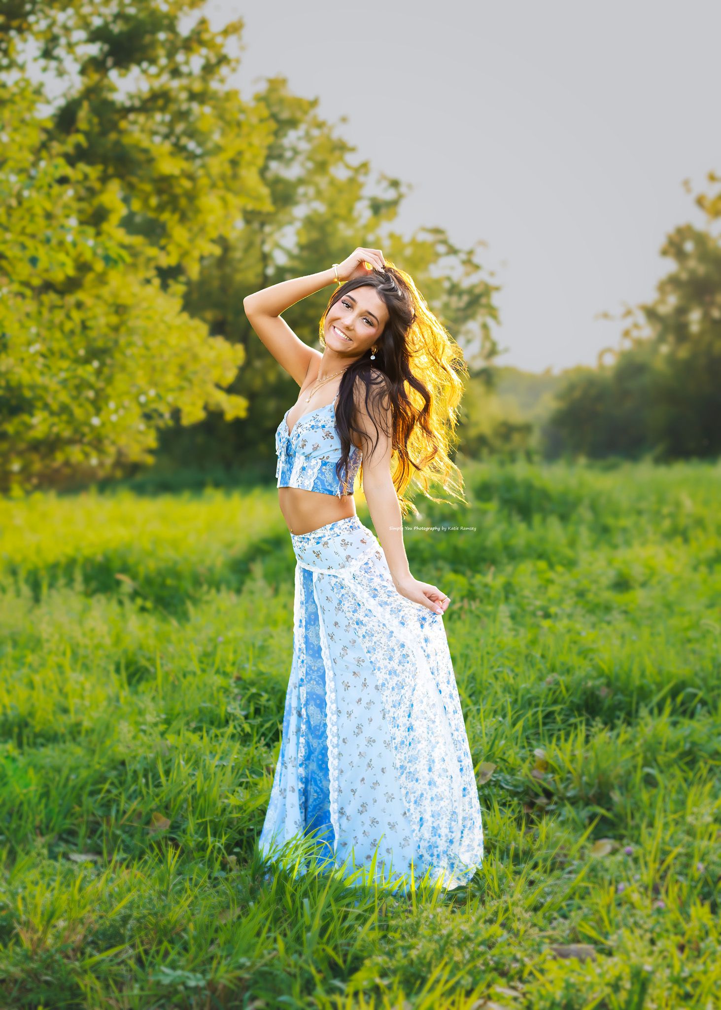 Young woman in blue floral crop top and maxi skirt posing in sunny green meadow with flowing hair.