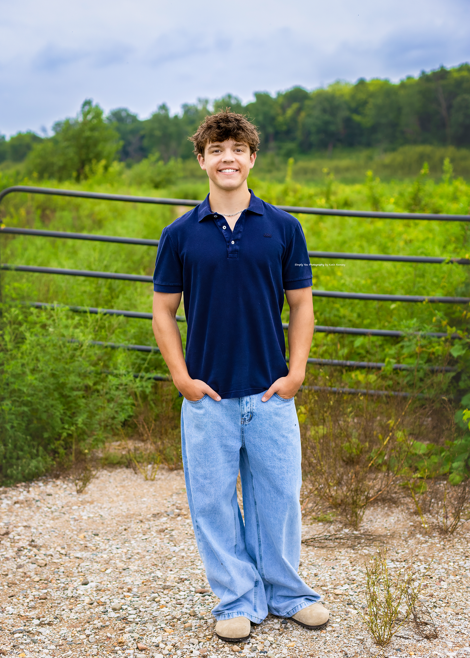Smiling teenage boy in navy polo shirt and light jeans standing on gravel path near metal gate with green foliage.