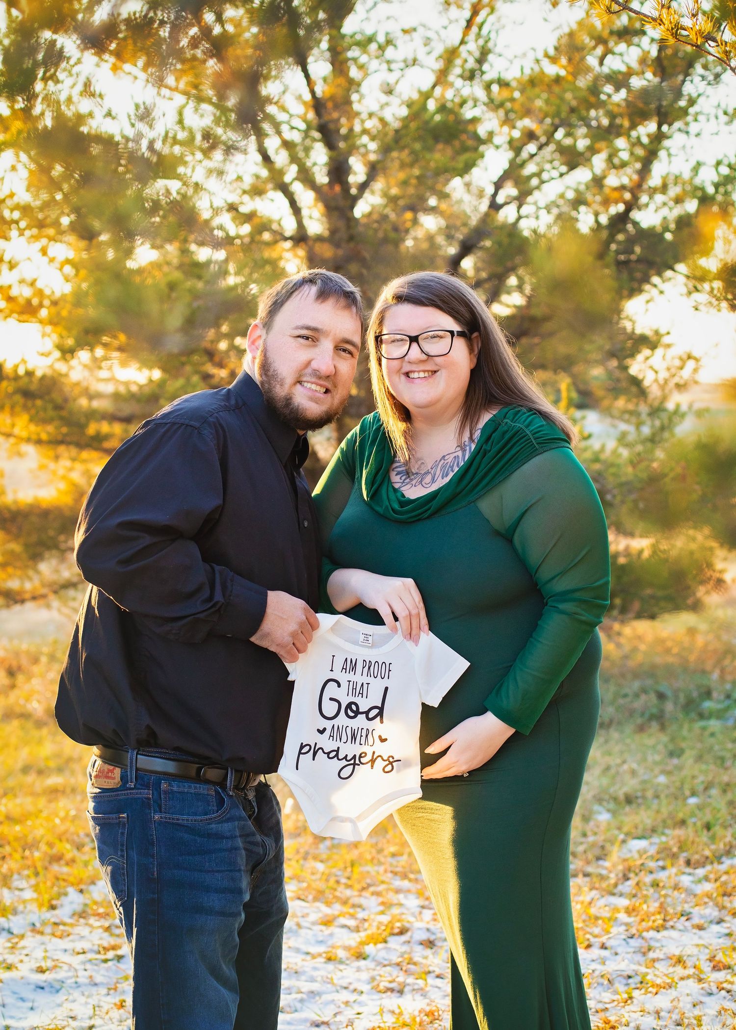 Smiling couple holding a God answers prayers baby onesie during maternity photo session.