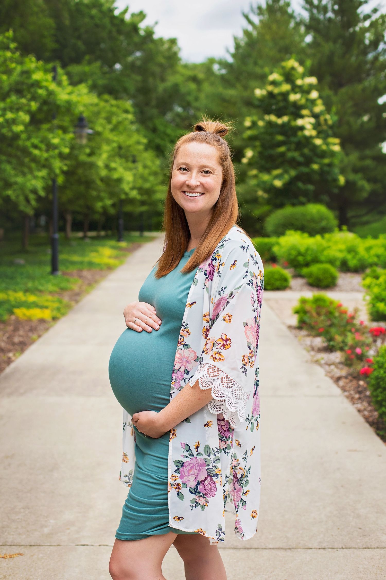 Glowing pregnant woman in teal dress and floral kimono cradles her baby bump while smiling on a garden path.