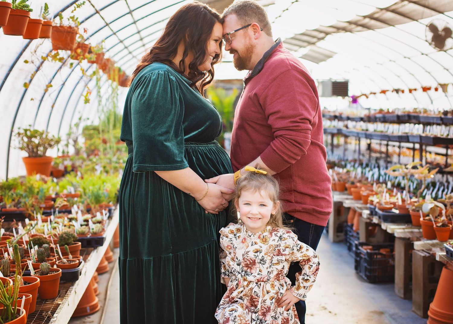 Expecting couple touches foreheads tenderly while toddler daughter smiles at camera in plant nursery.
