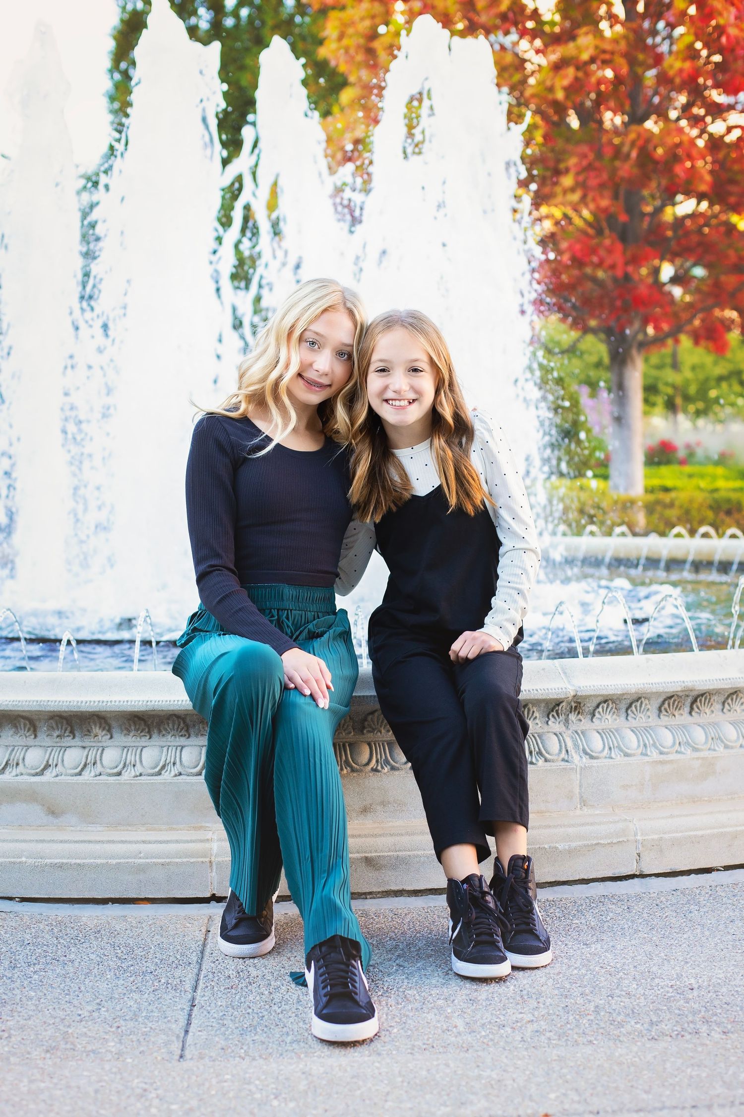 Two girls sitting together by a fountain with colorful autumn trees behind them.