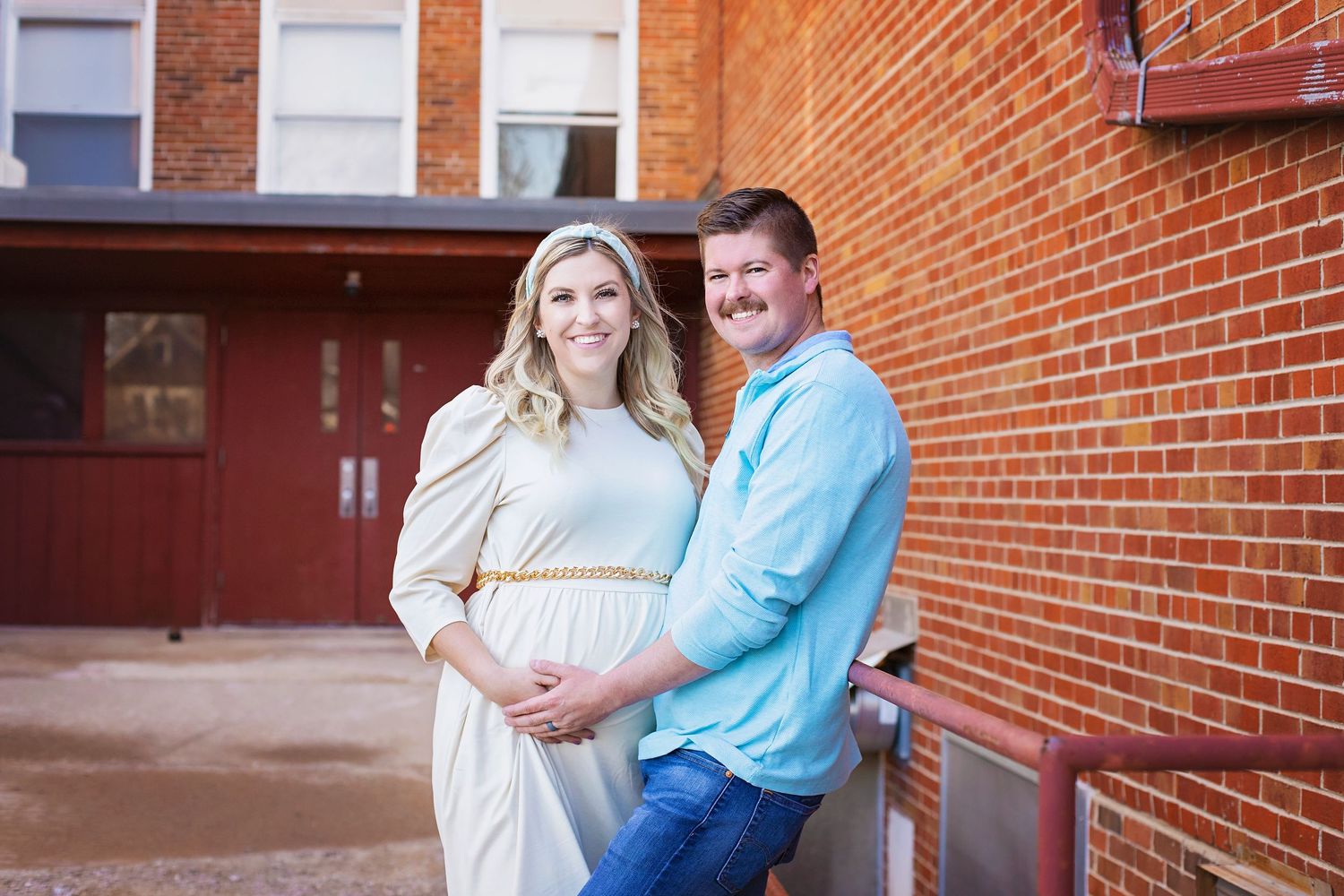 Happy expecting couple posing outdoors during maternity photo session by brick building.