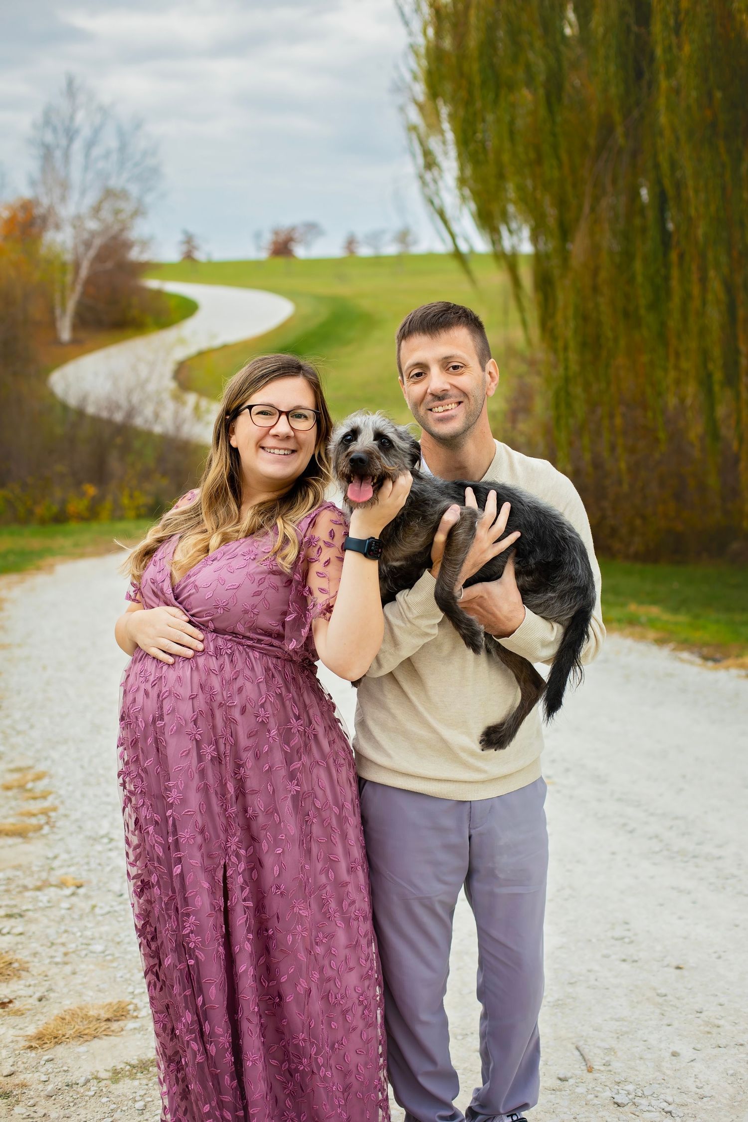 Pregnant woman in purple lace gown and man holding black dog pose on scenic winding gravel path.