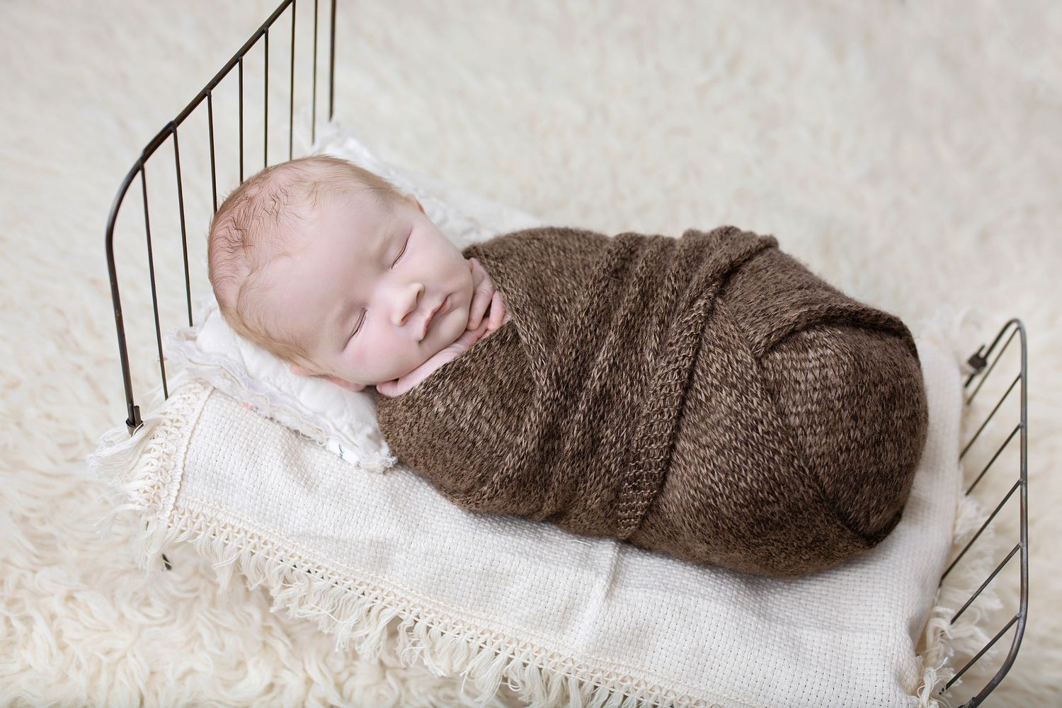 Smiling newborn baby swaddled in a brown knit wrap lying in a small vintage metal bed on white fur.