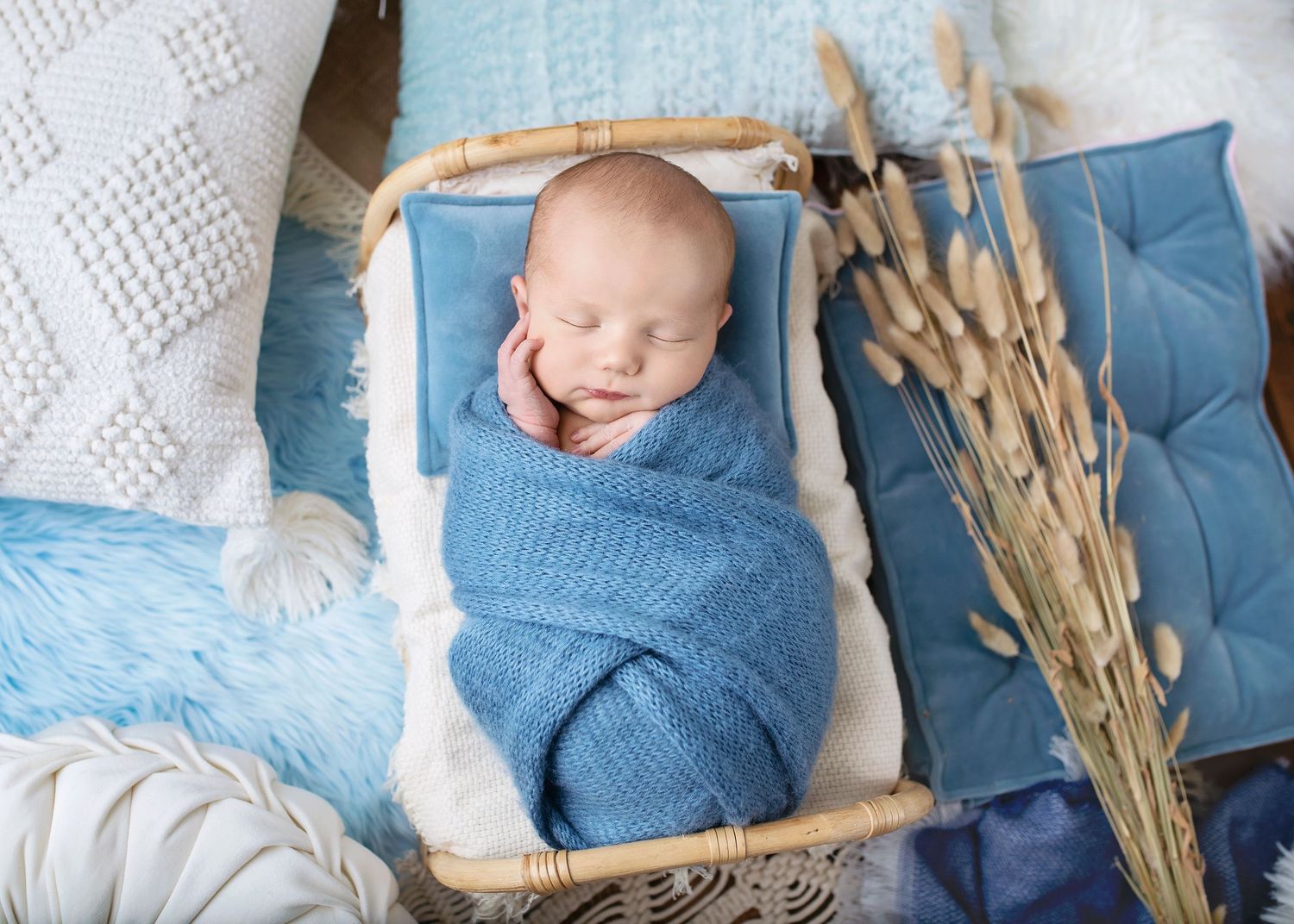 Newborn baby swaddled in blue knit wrap sleeping in a wicker bassinet with boho decor.