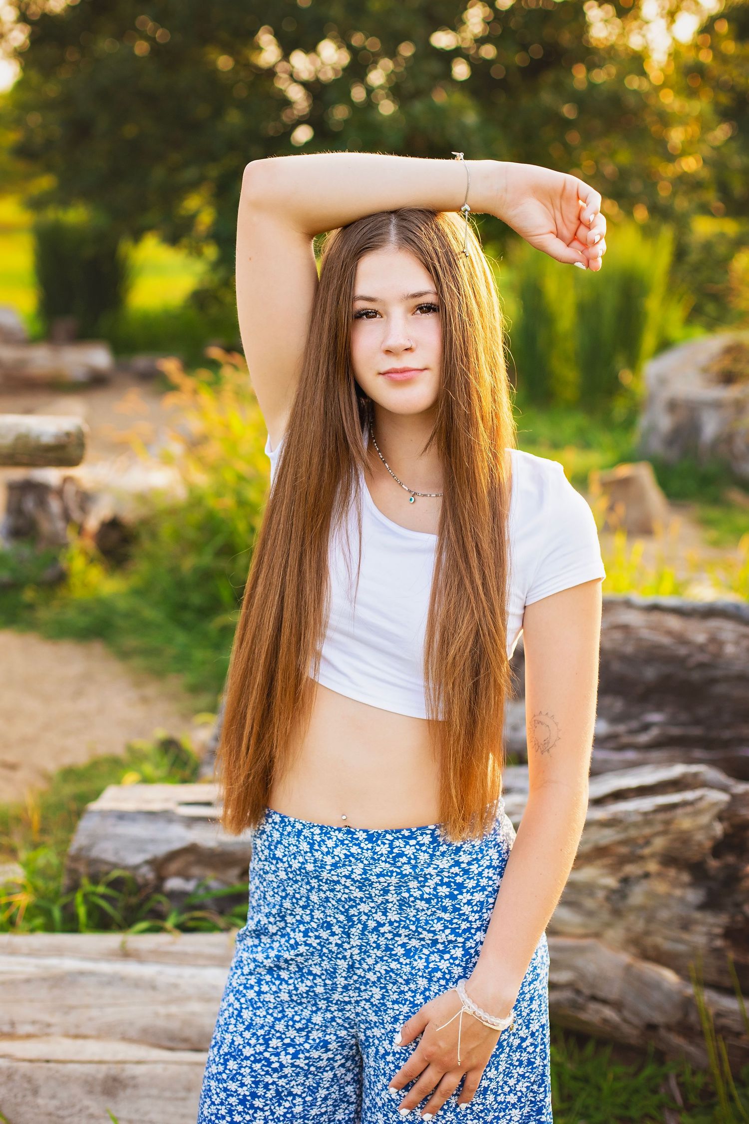 Teen girl with long hair in white crop top and floral pants posing outdoors in golden sunlight.