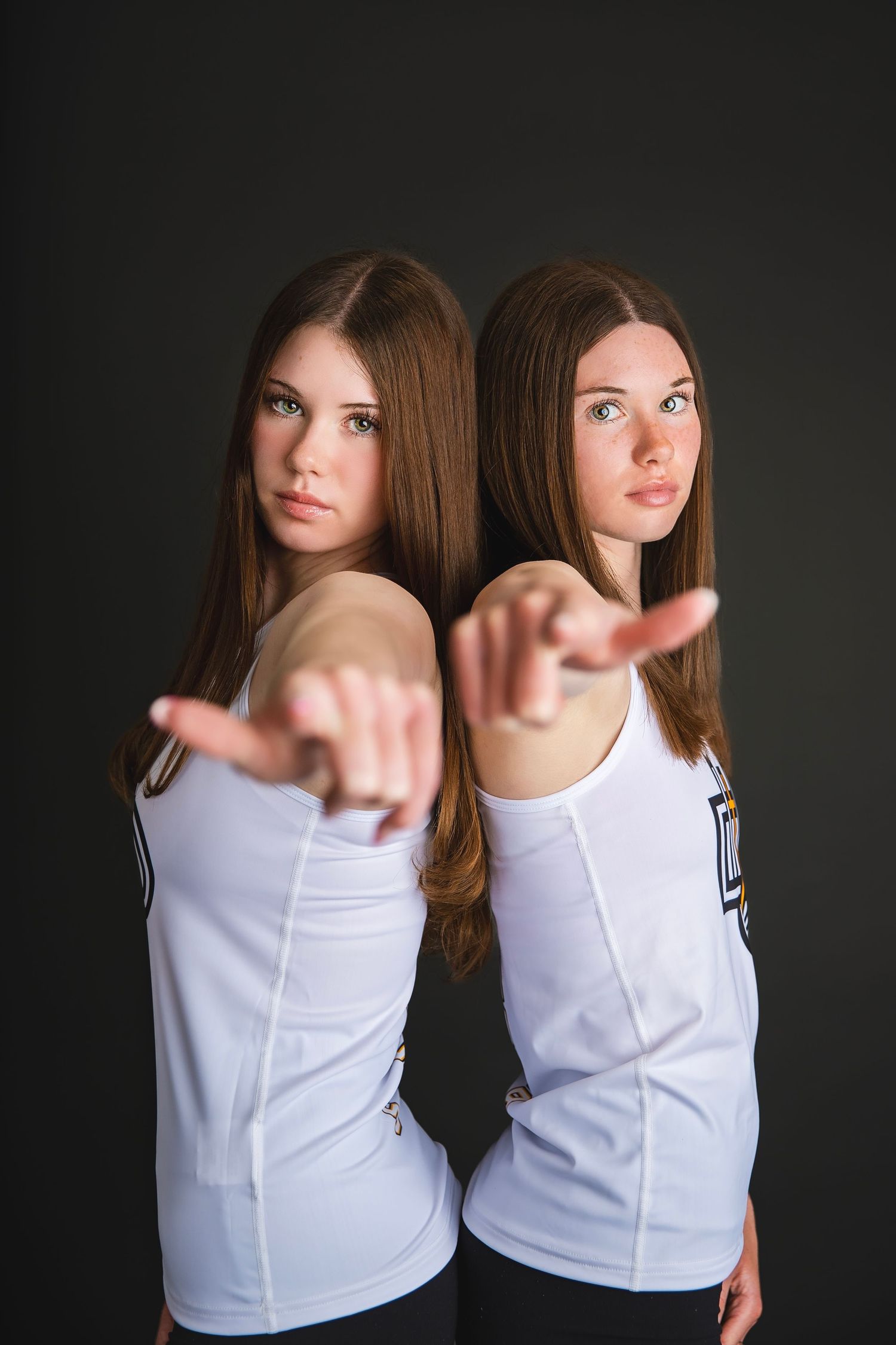 Two teenage girls in white volleyball uniforms pointing at camera against a dark studio background.