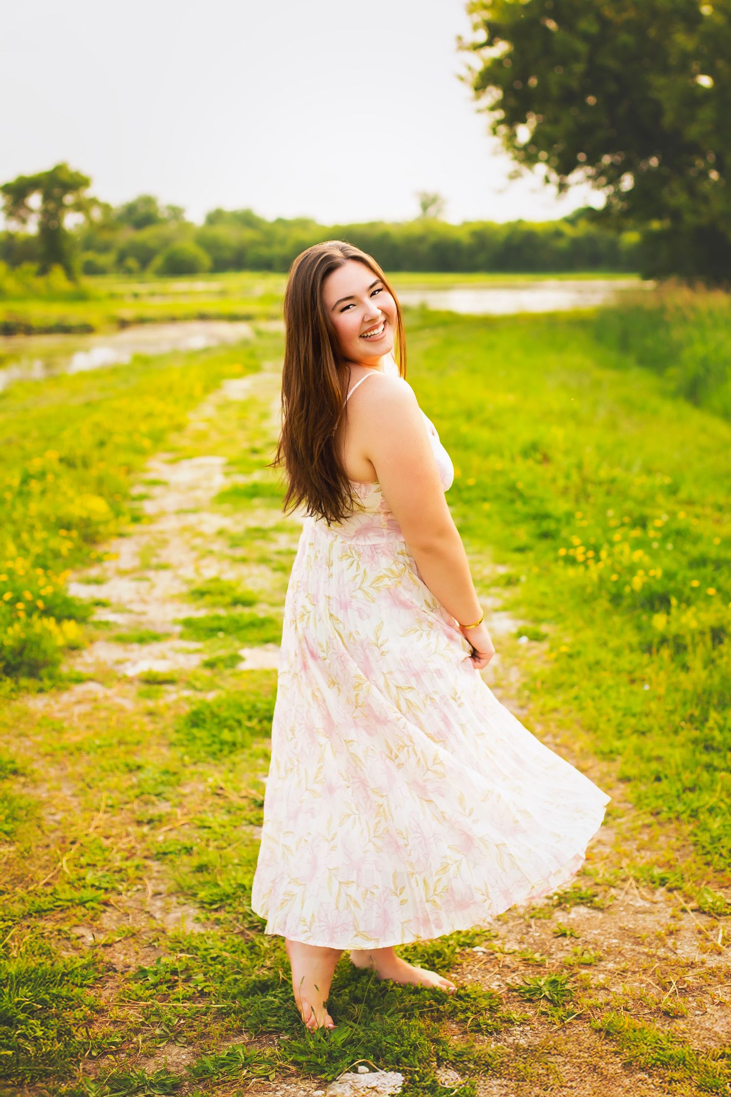 Laughing young woman in floral dress twirling on grassy path near river.