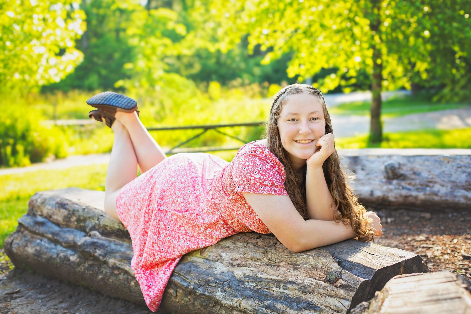 Smiling girl in pink floral dress lying on wooden log in sunny green park setting.