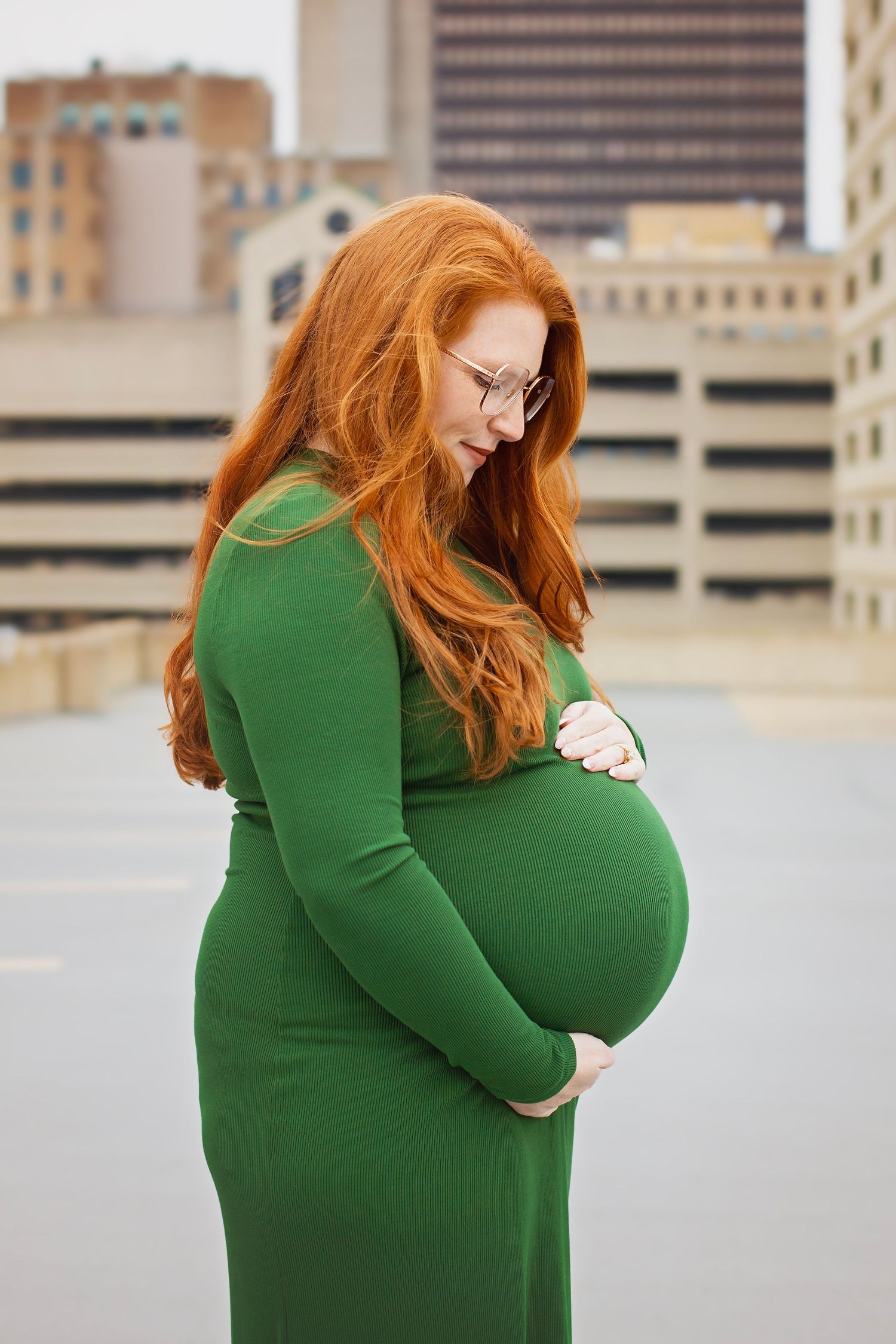 Redheaded pregnant woman in green dress lovingly looks down at her baby bump.
