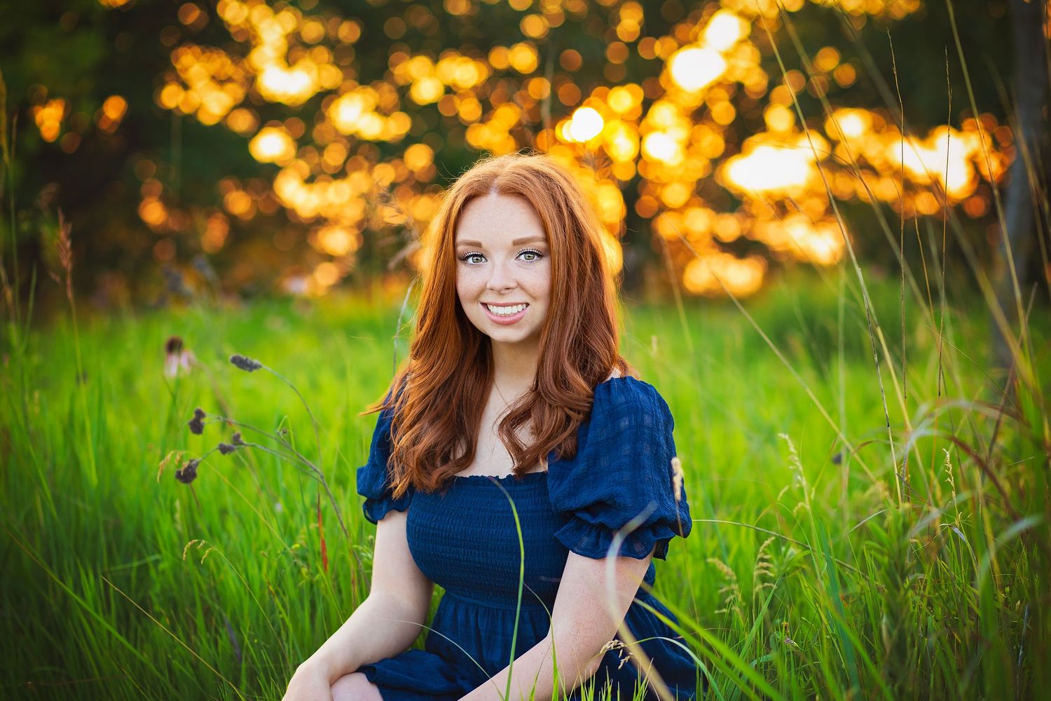 Smiling redhead teen girl in navy blue dress sits in green meadow with golden sunset bokeh background.