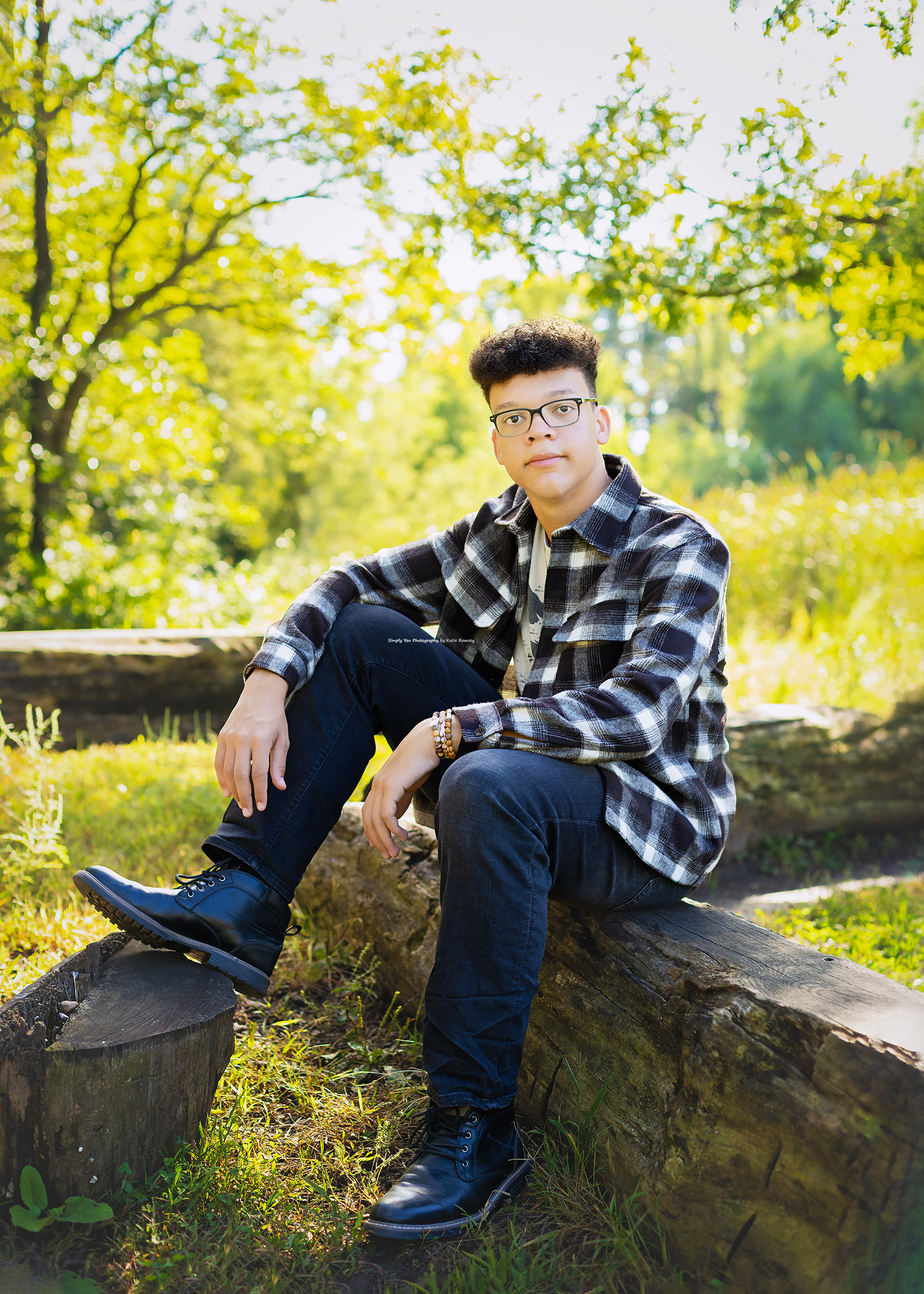 Young man in plaid shirt and glasses sitting on stone wall outdoors in lush green park setting.