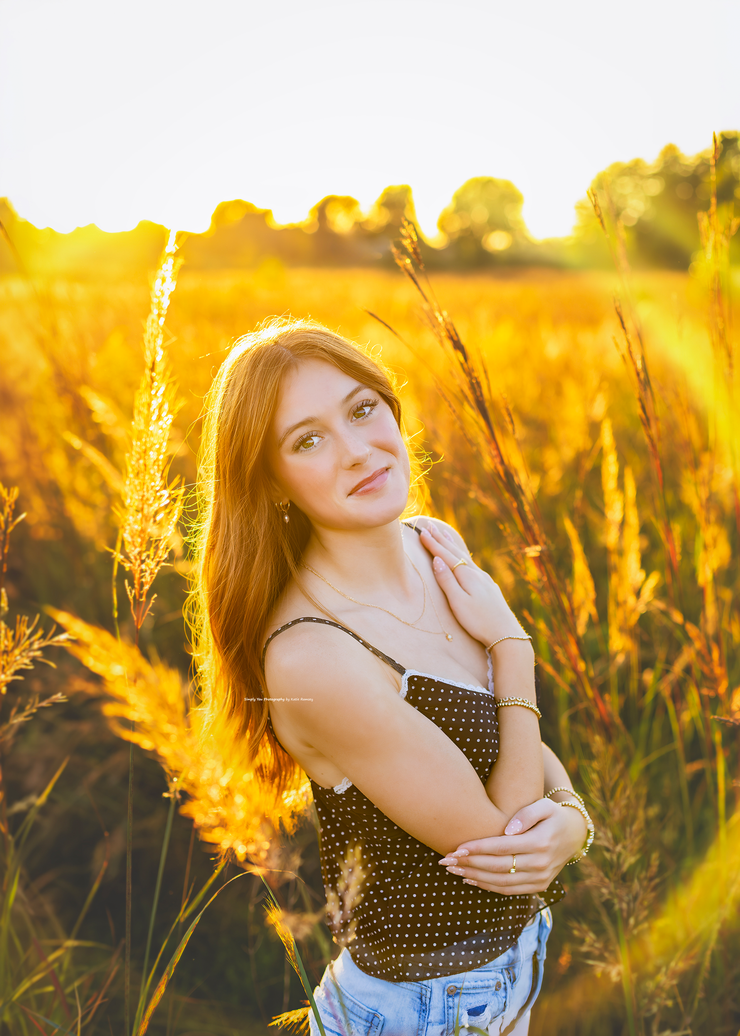 Young woman in polka dot top poses in golden wheat field during sunset, warm light glowing behind her.