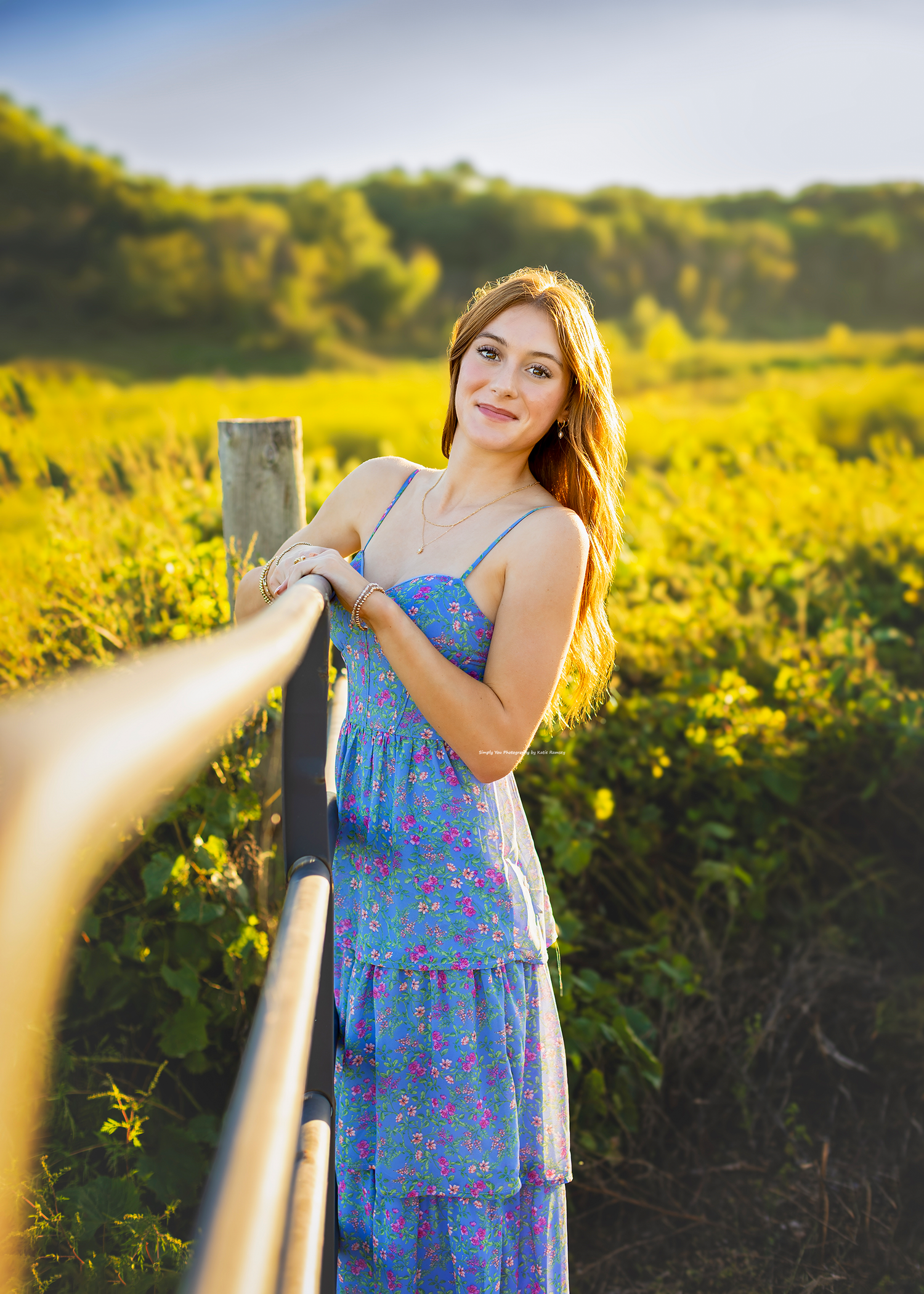 Young woman in floral blue dress leaning on wooden fence in sunlit green meadow, smiling warmly at camera.