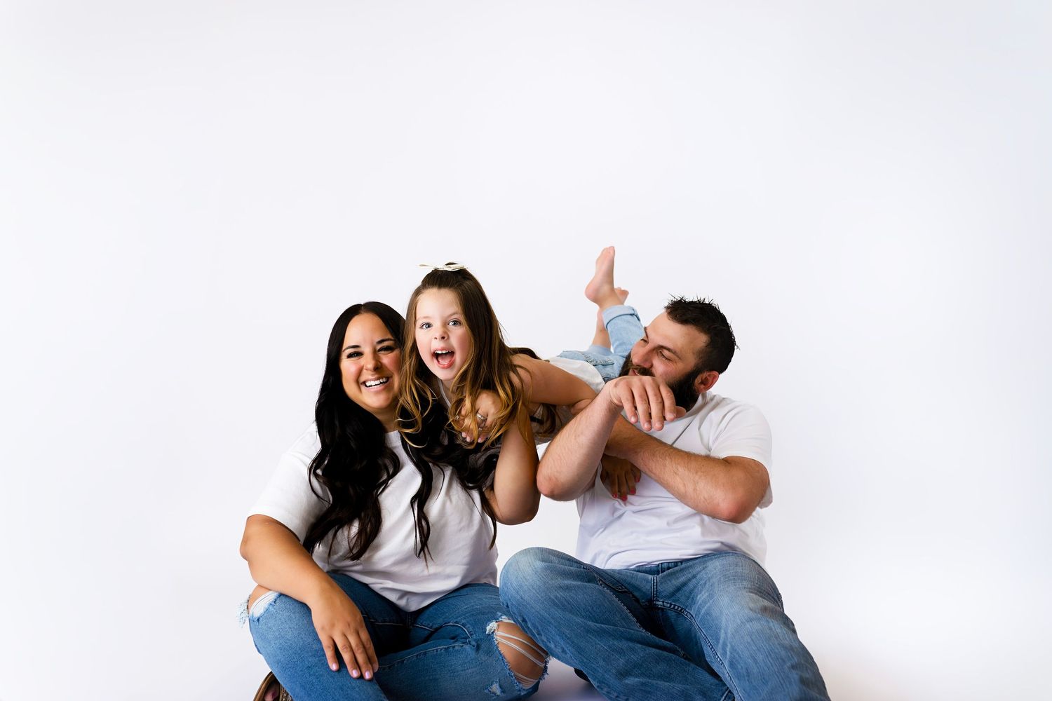 A family of three in white tees and jeans laugh together as parents playfully lift their young daughter on a white backdrop.