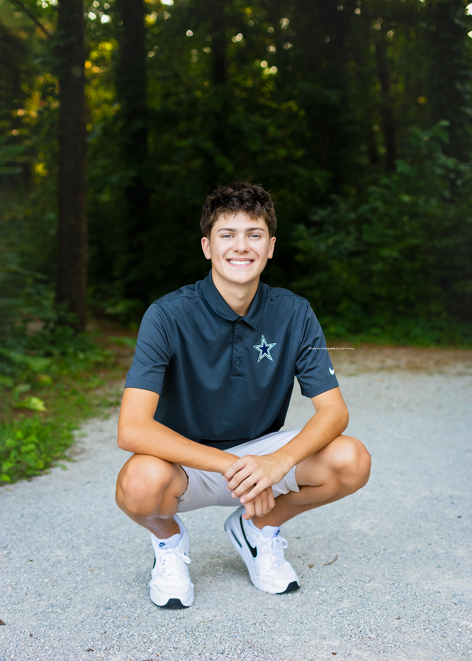 Smiling young man crouching outdoors in a navy Nike polo shirt and white shorts, posing on a gravel path with trees behind.