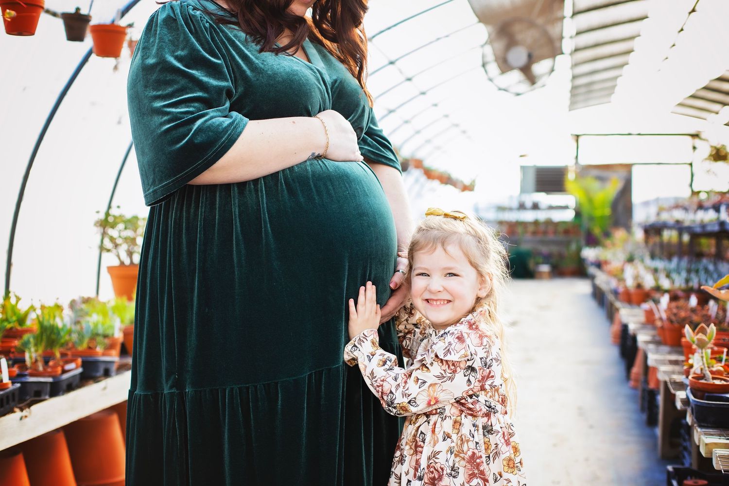 Smiling toddler girl in floral dress gently touches pregnant mothers belly in a sunny greenhouse nursery.
