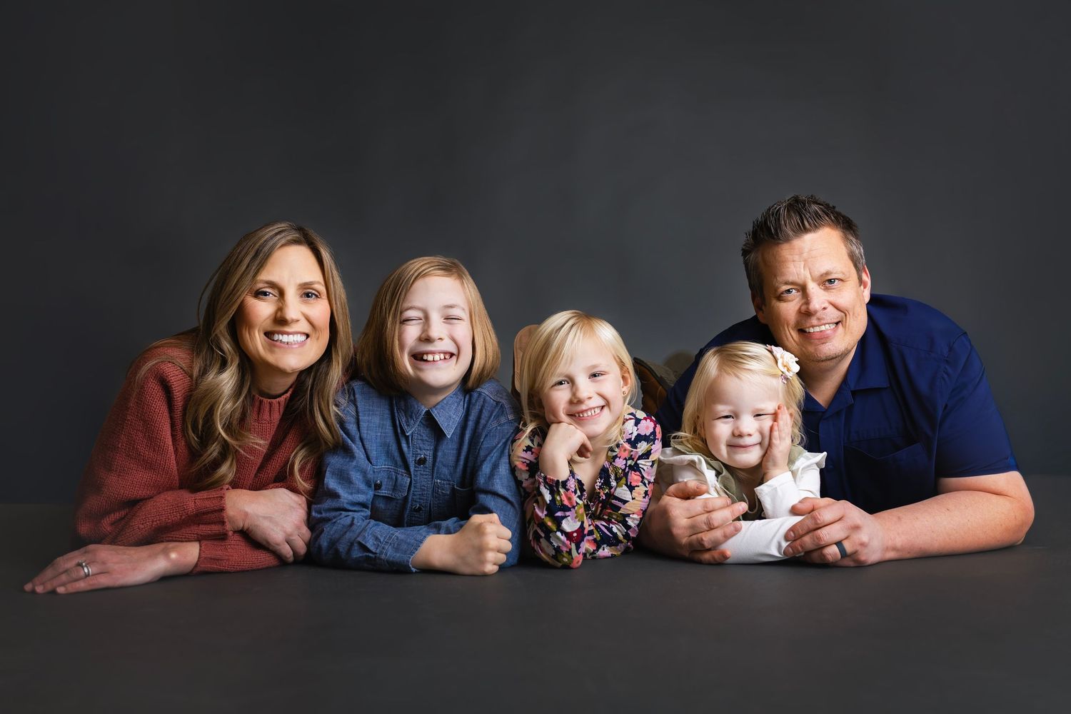 Cheerful family of five lying together and smiling at camera against a dark gray background.