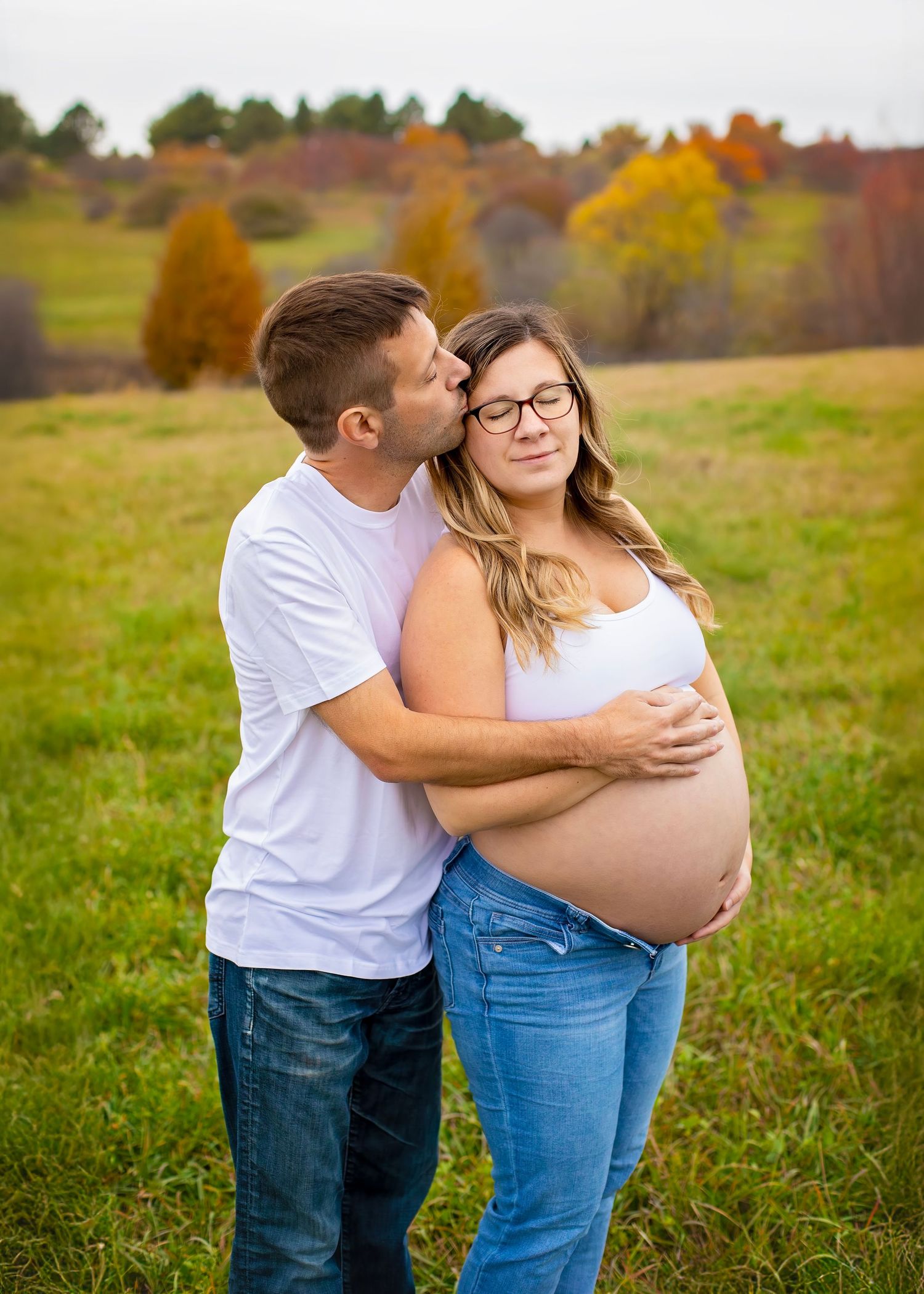 Man in white shirt tenderly kisses pregnant woman's temple while embracing her in green field.