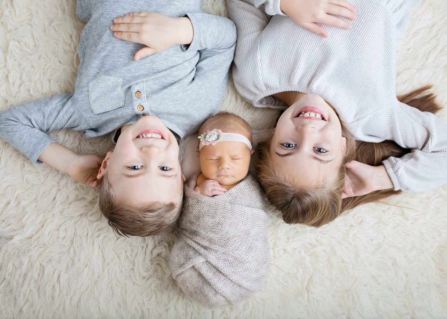 Three siblings lying on white fur rug with newborn baby wrapped in gray swaddle in the middle.