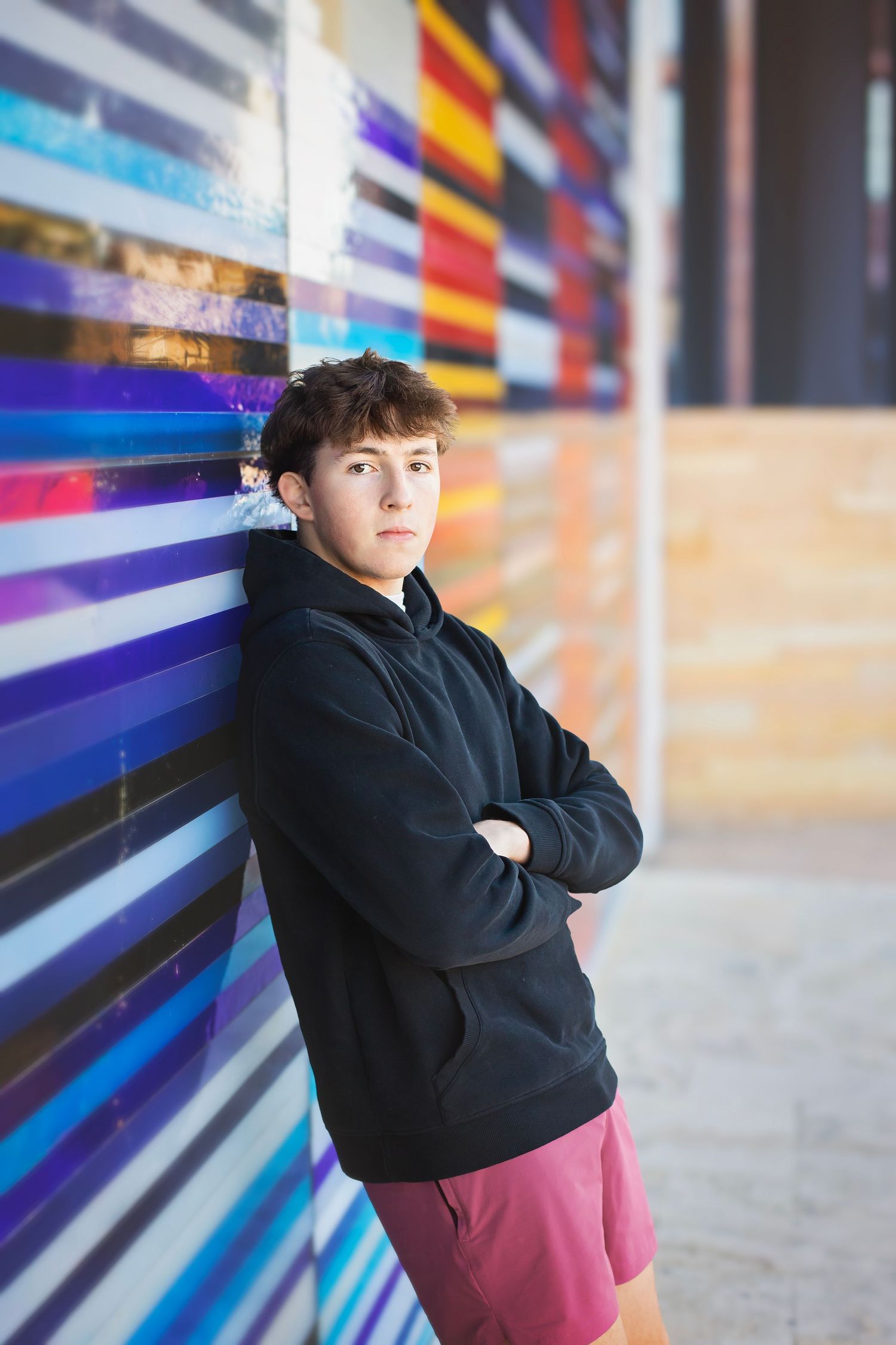 Teen boy in black hoodie leans against colorful striped wall with arms crossed.