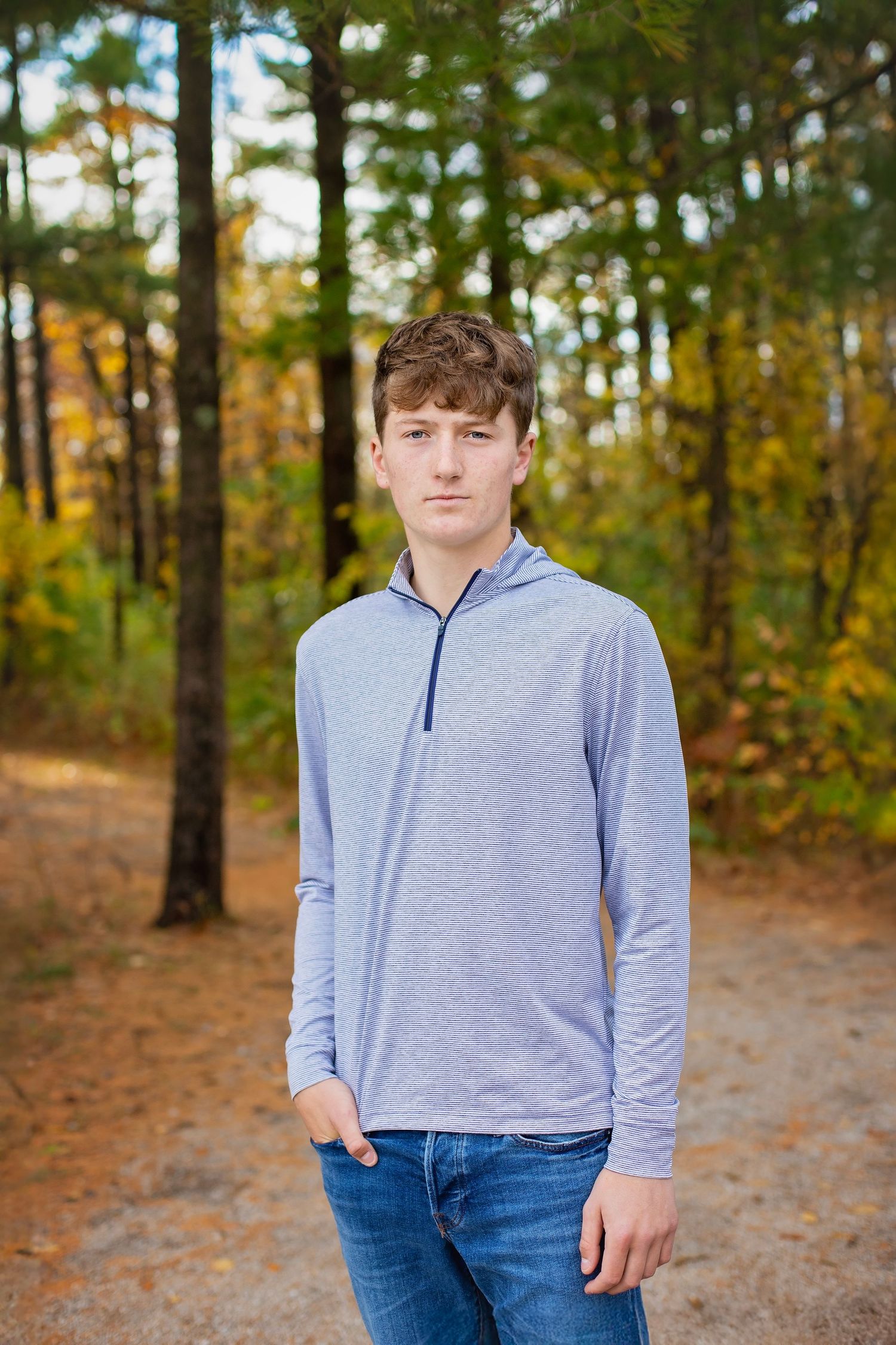 Teen boy in light blue quarter-zip hoodie standing on a forest path surrounded by fall foliage.