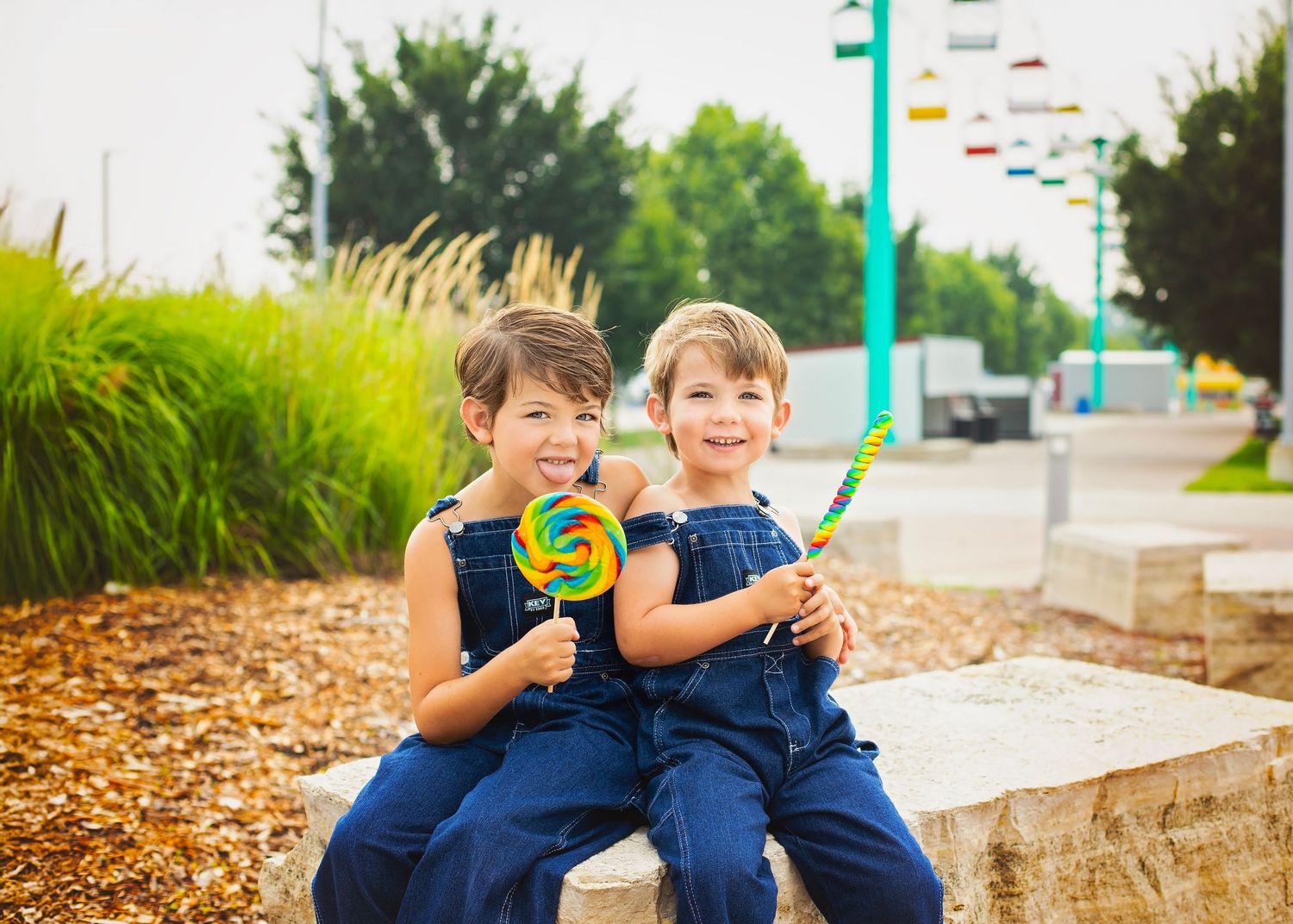 Twin boys in matching denim overalls smile holding large colorful lollipops outdoors in summer.
