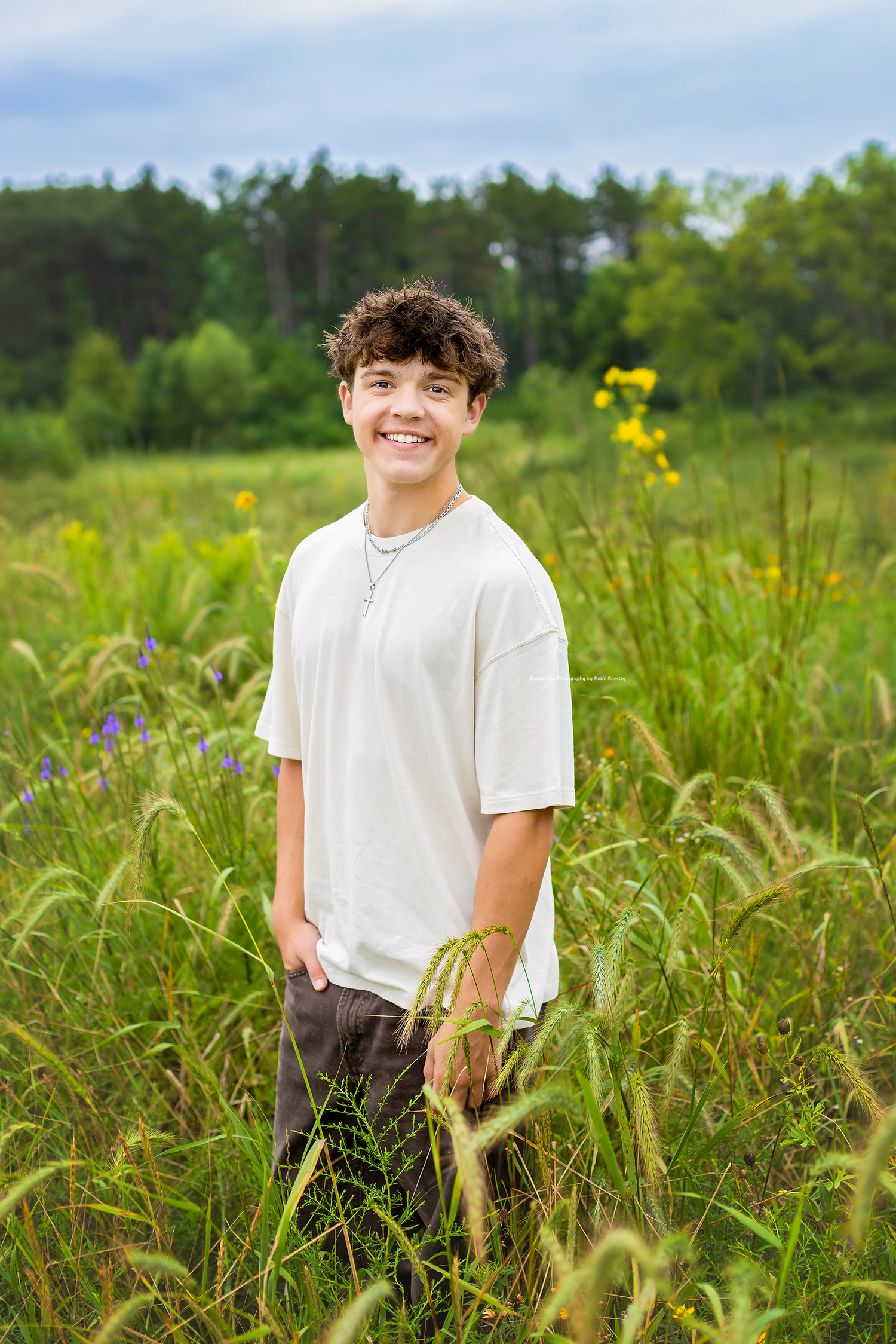 Smiling teenage boy in white t-shirt standing in a lush green meadow with wildflowers and trees in background.