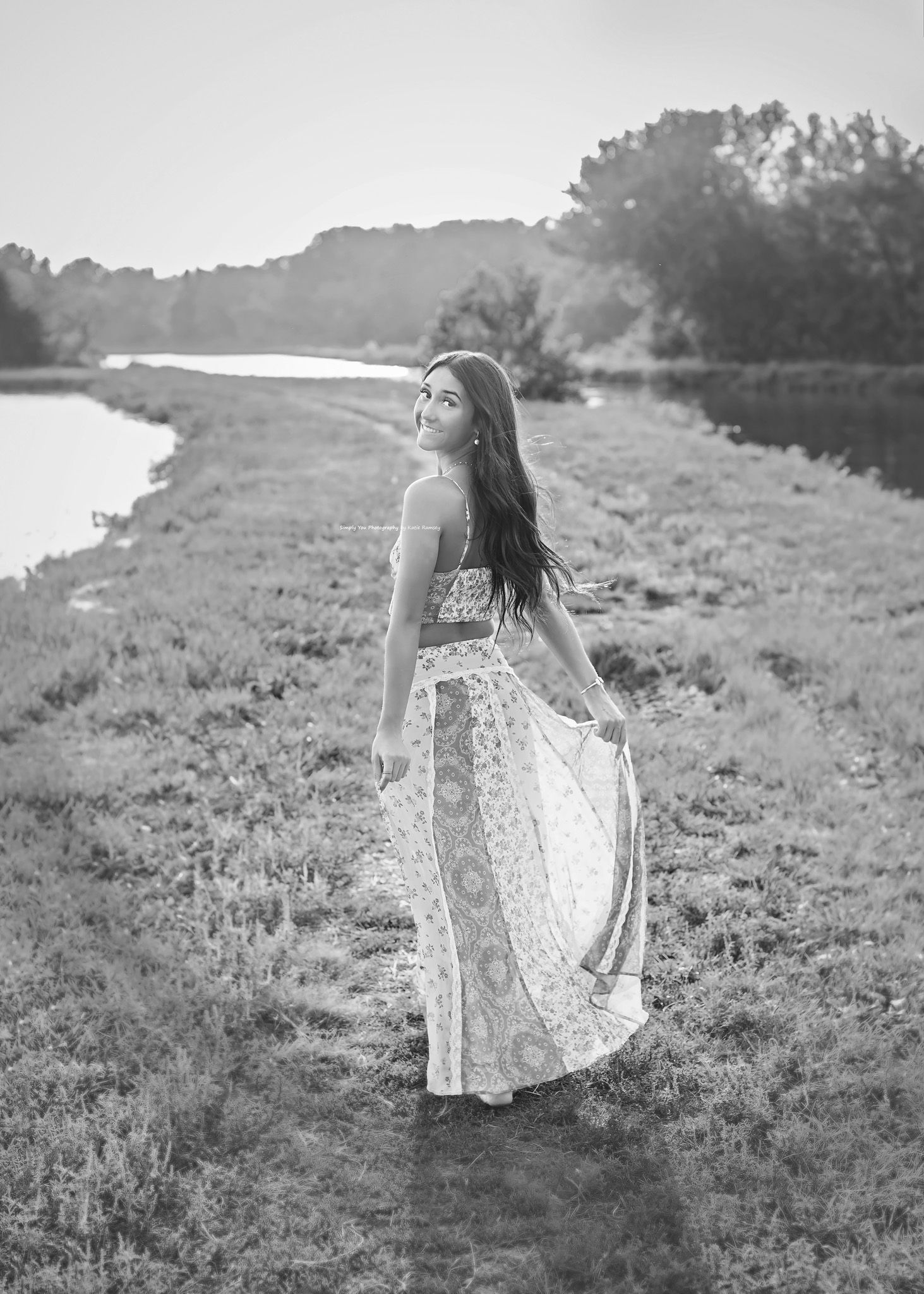 Young woman in boho floral maxi dress walking along scenic riverside path, elegant black and white portrait.