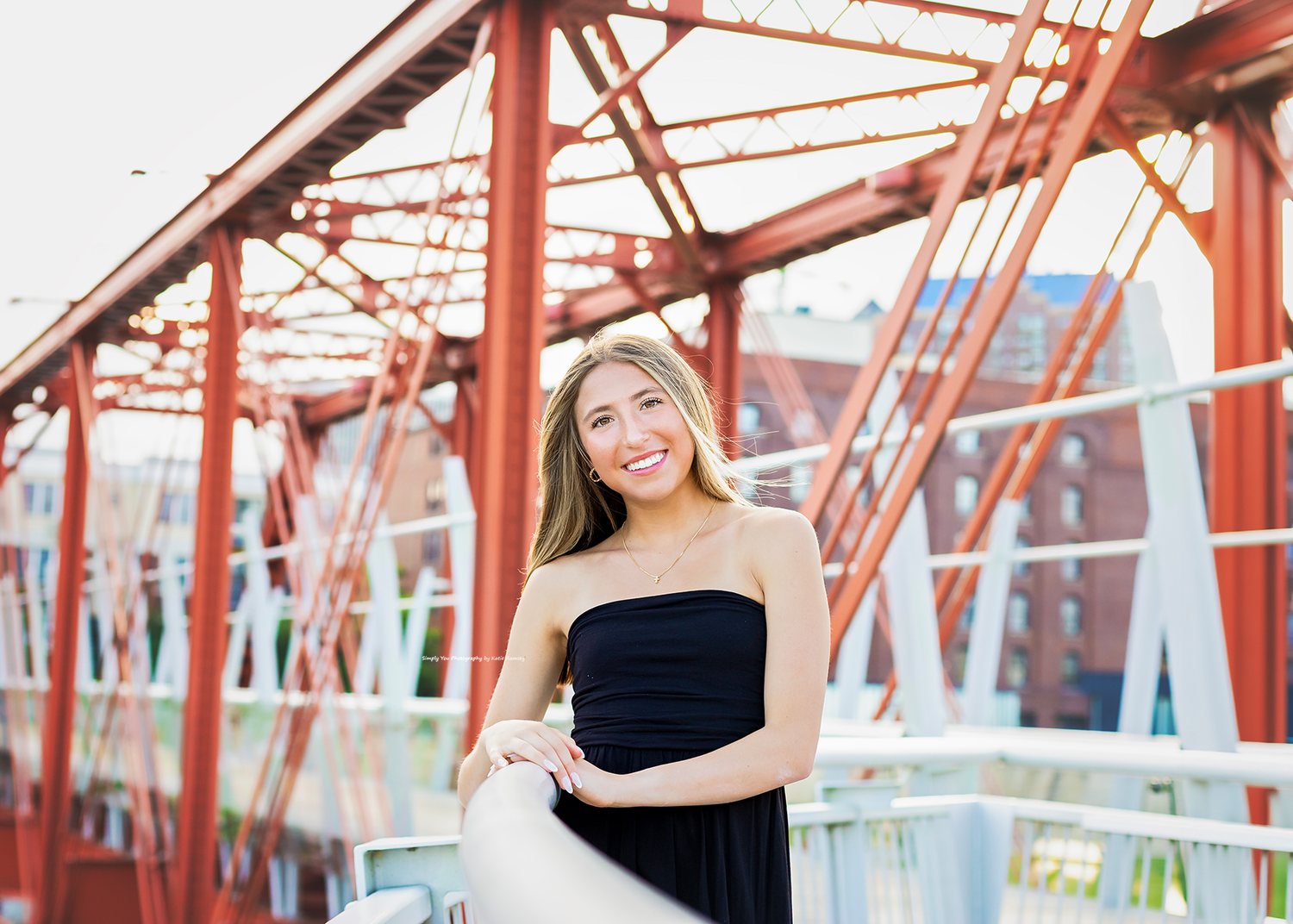 Young woman in black strapless dress smiling on a red steel bridge with urban background.