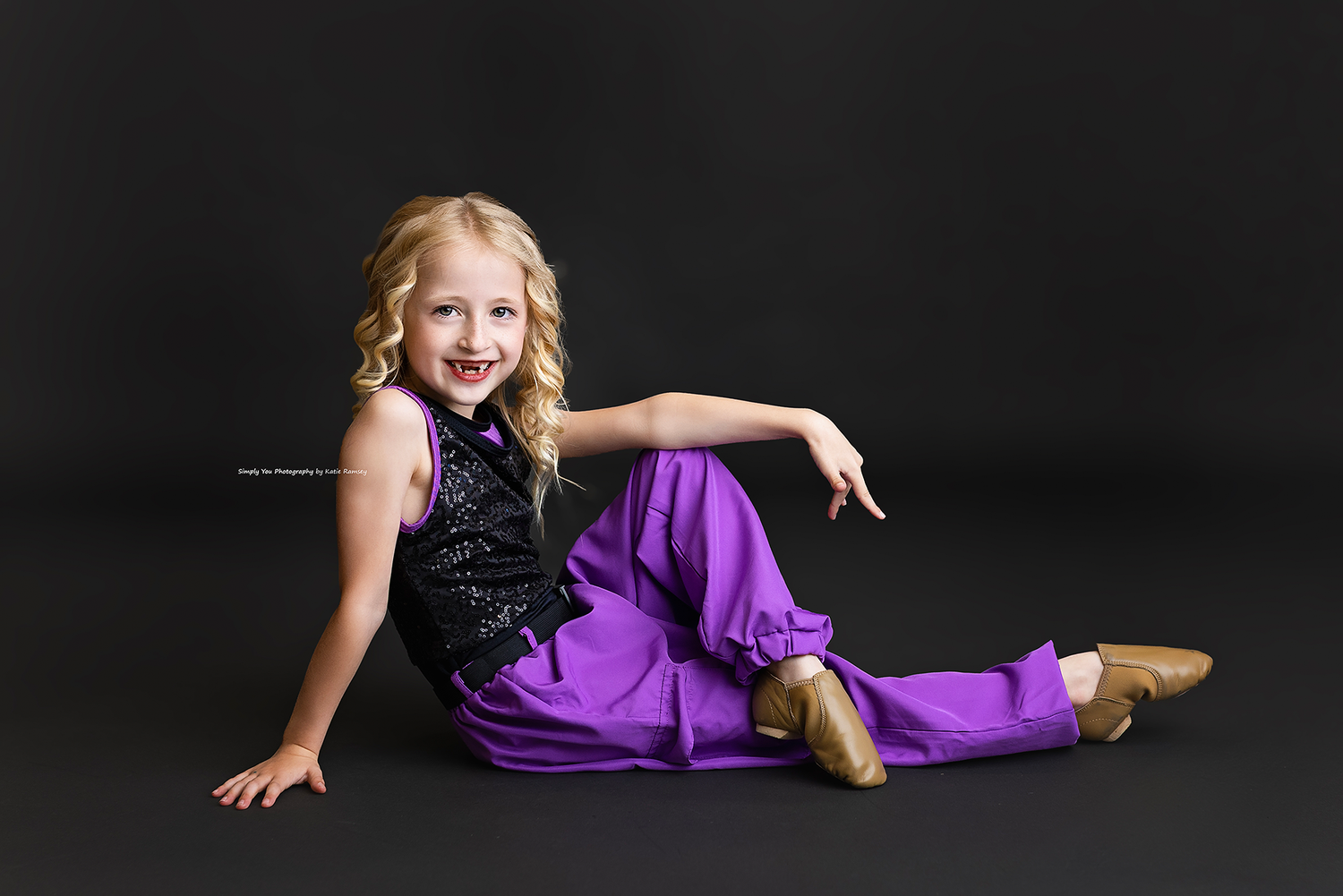 Young girl in purple pants and black sequin top poses gracefully on dark studio floor wearing gold dance shoes.
