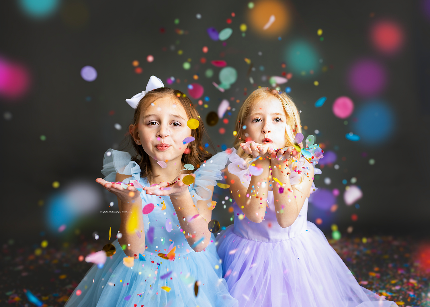Two girls in princess dresses blowing colorful confetti toward camera in studio setting.
