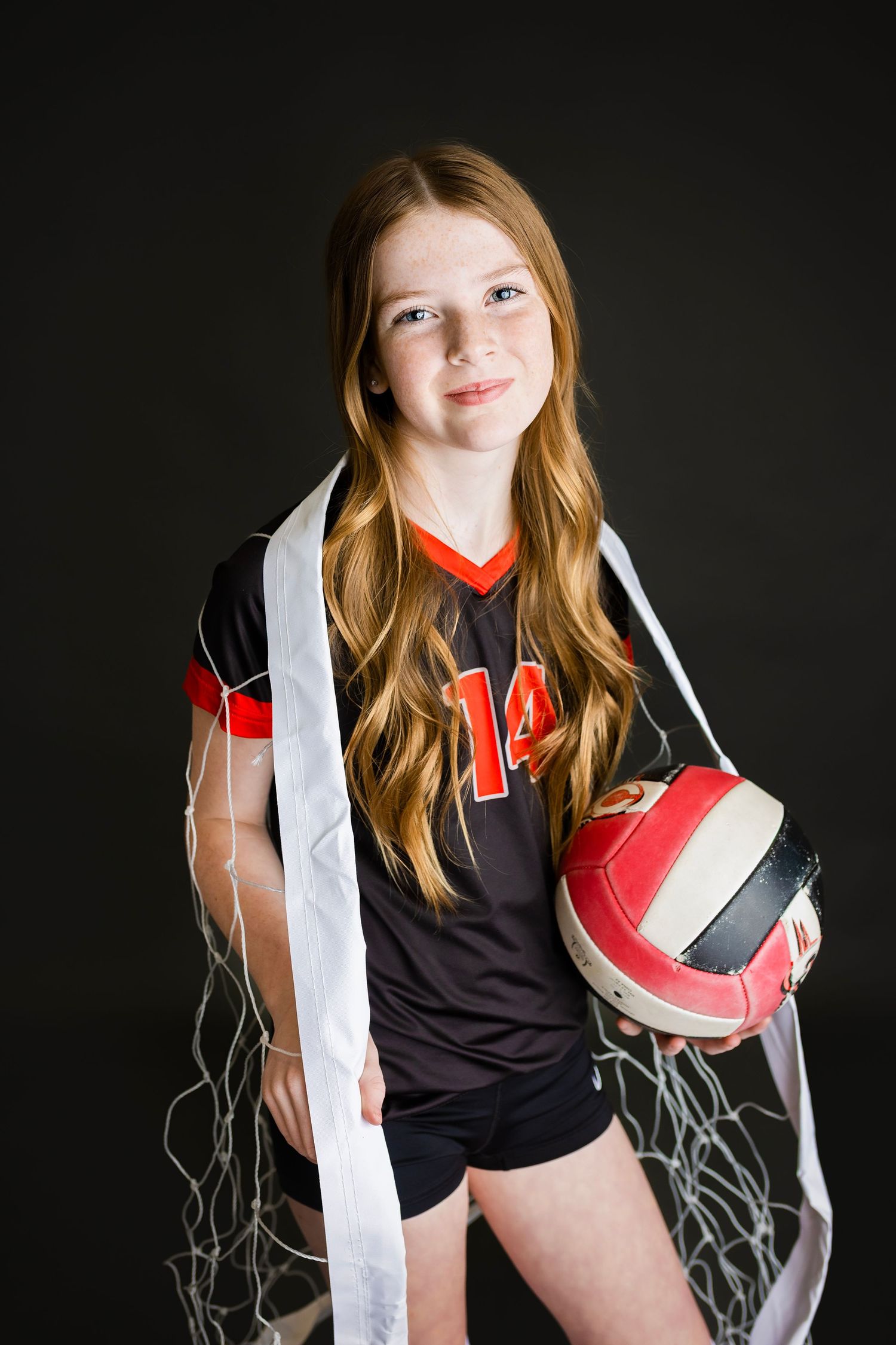 Young girl wearing number 14 volleyball jersey holds a Baden ball while draped in a volleyball net on dark background.