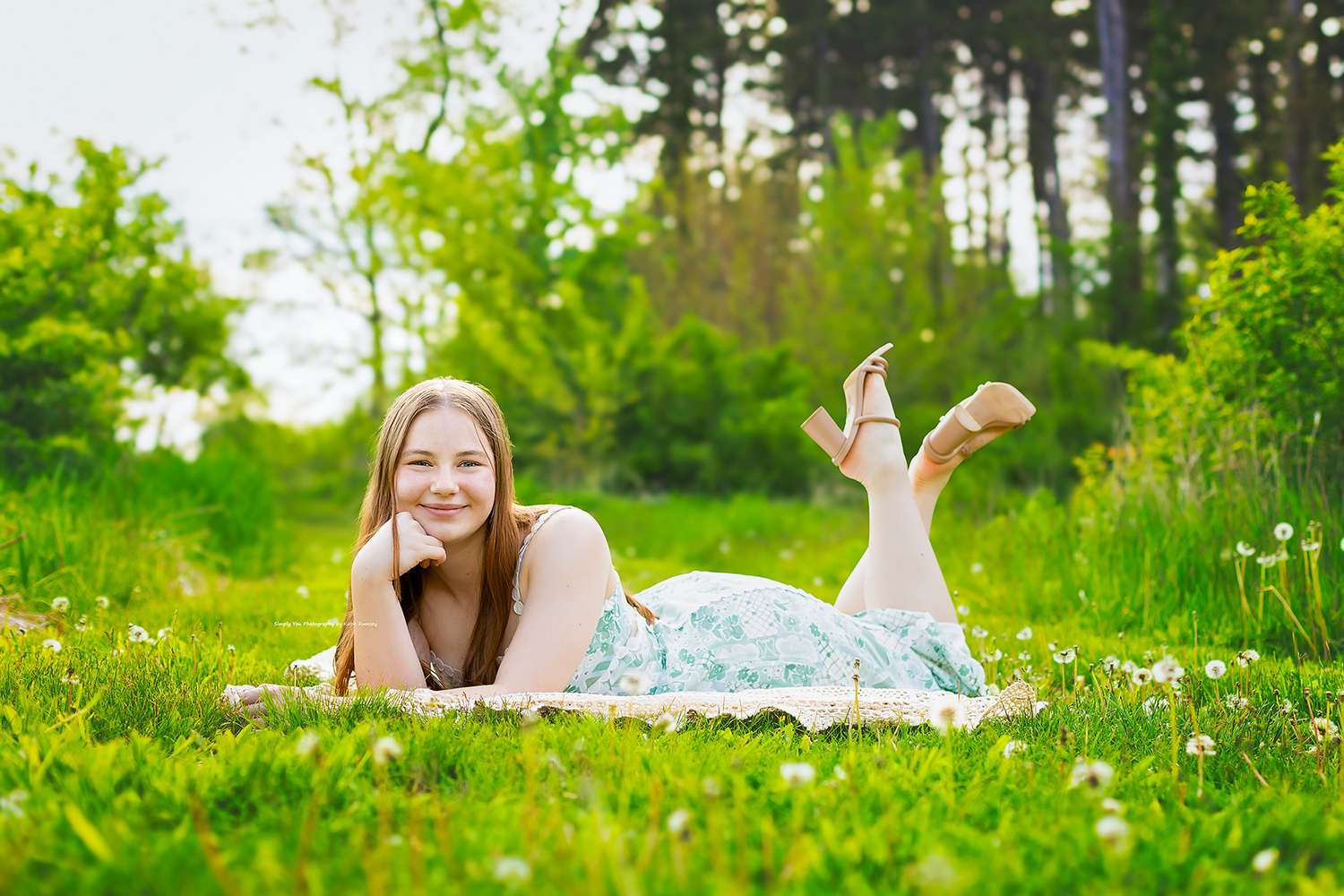 Young woman in floral dress lying on grass in sunny green park, smiling cheerfully during outdoor portrait session.