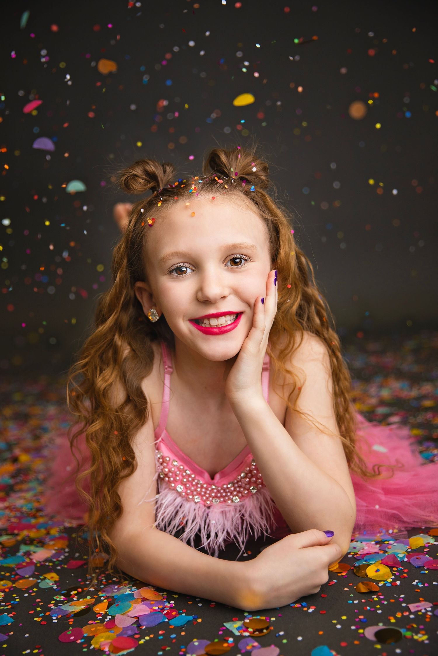 Smiling girl in pink feathered dress lying on confetti-covered floor with hand on cheek looking at camera.