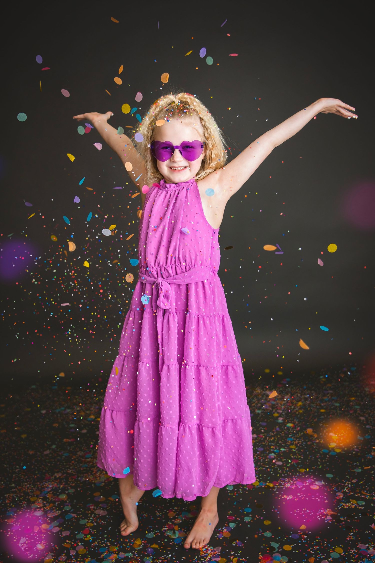 Joyful girl in pink dress and sunglasses dancing with colorful confetti on dark background.