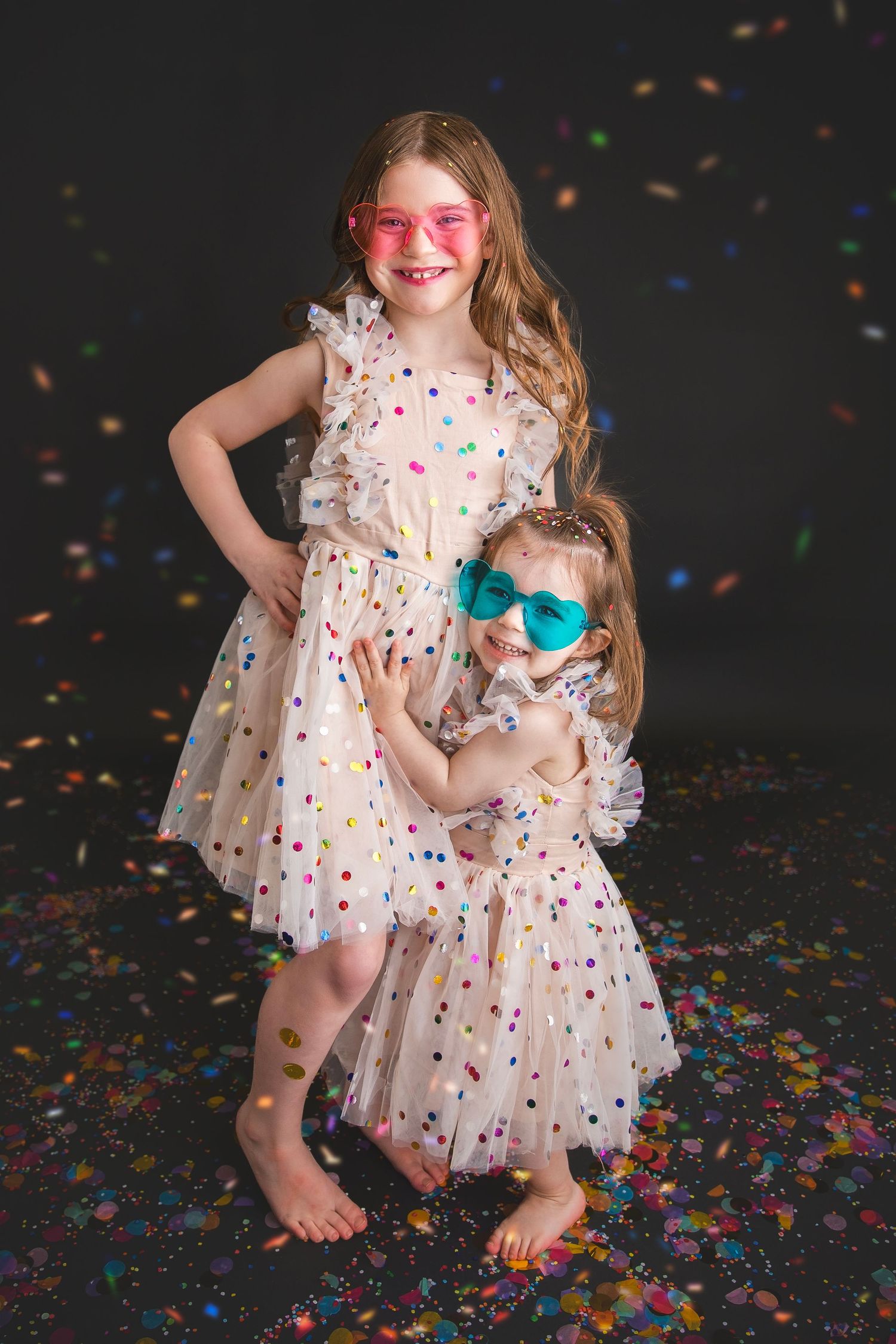 Two sisters in polka dot dresses wearing heart sunglasses with falling confetti.