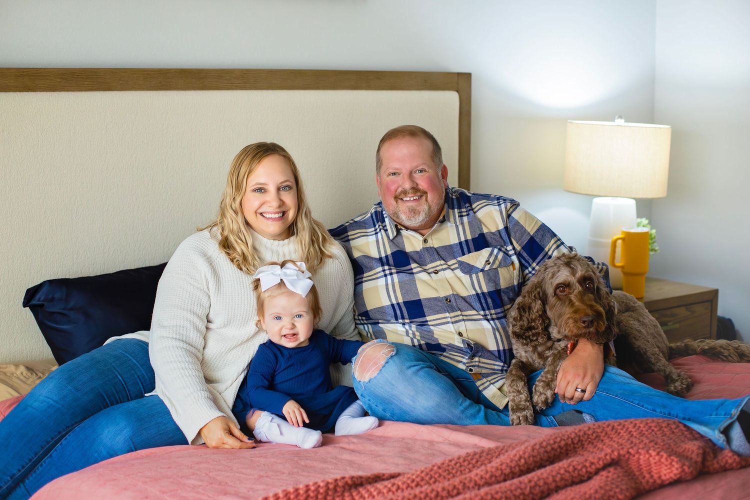 Smiling family with baby girl and brown dog sitting together on a bed in a cozy bedroom.