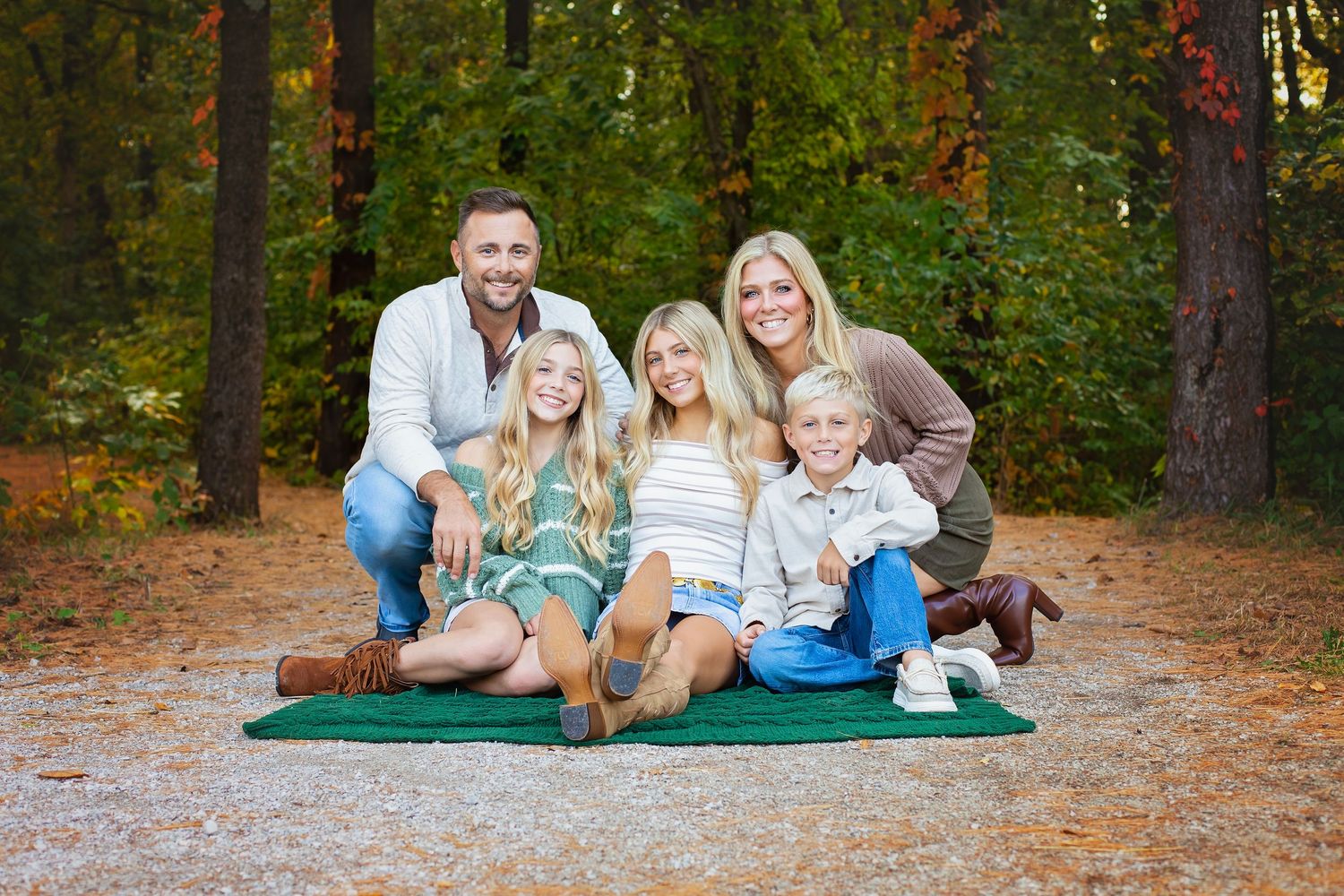 A smiling family of five poses together on a green blanket on a scenic autumn forest path.