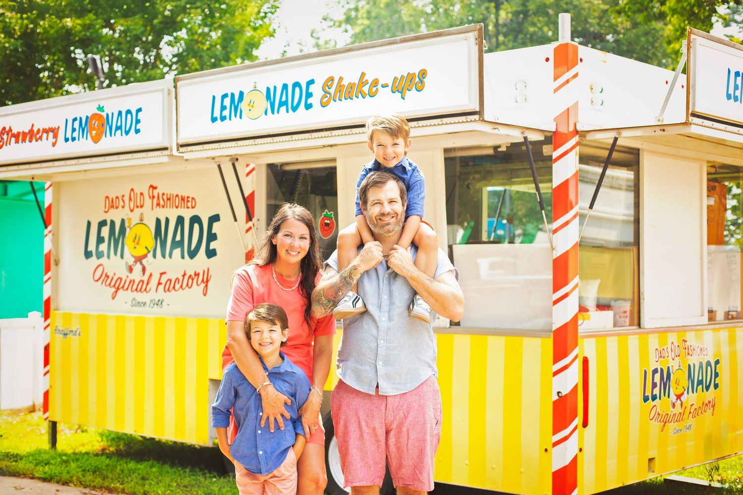 Happy family of four posing in front of a lemonade shake-up food stand at a summer fair.