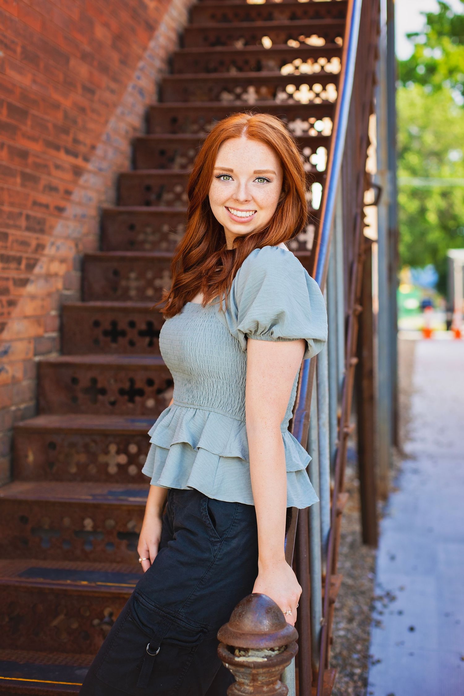 Happy redhead teen girl in sage top leans against railing of decorative brick staircase, smiling brightly.